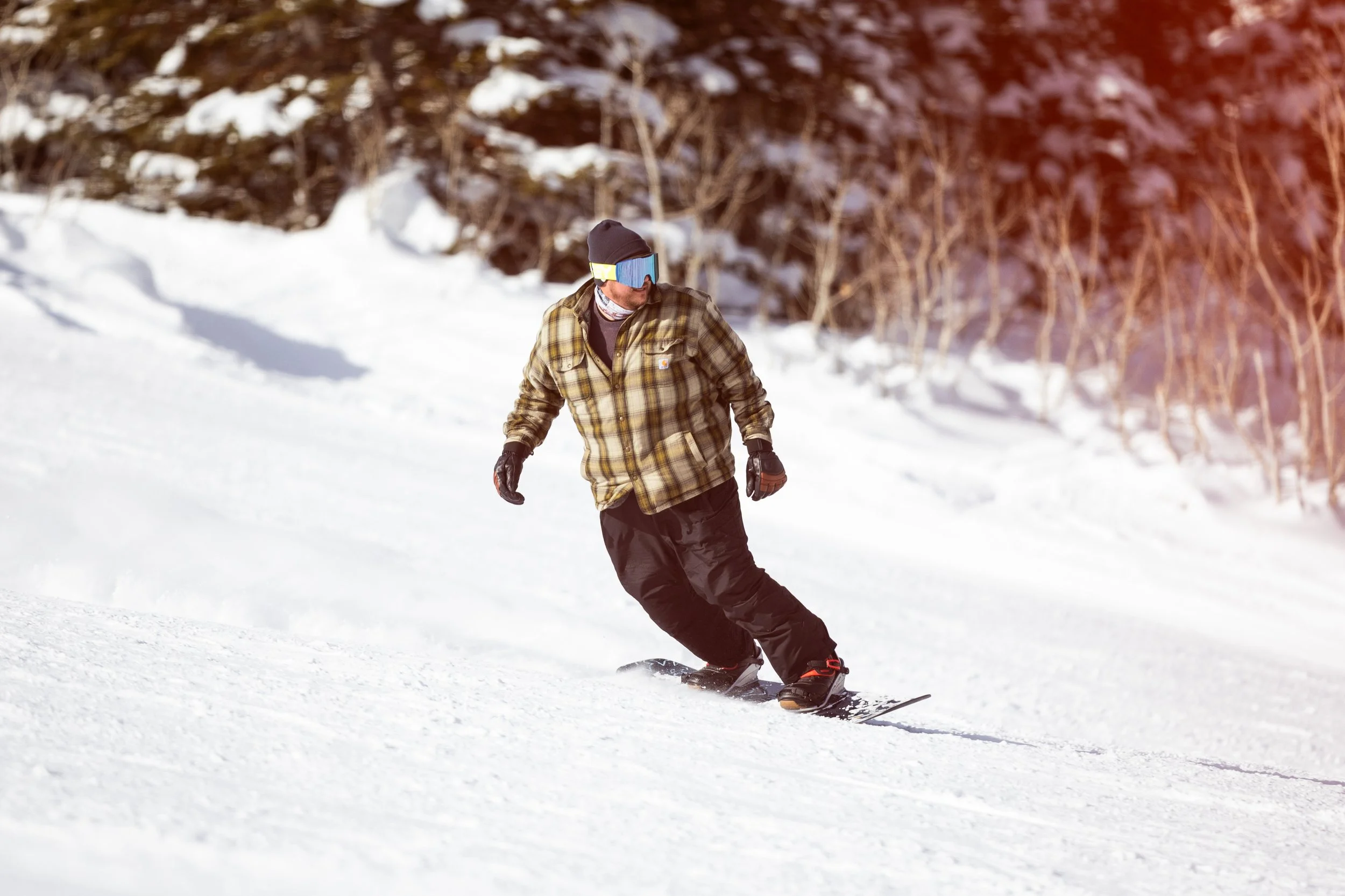 A man snowboarding on a snowy slope with a wooded background, wearing a plaid jacket, black snowpants, gloves, and goggles.