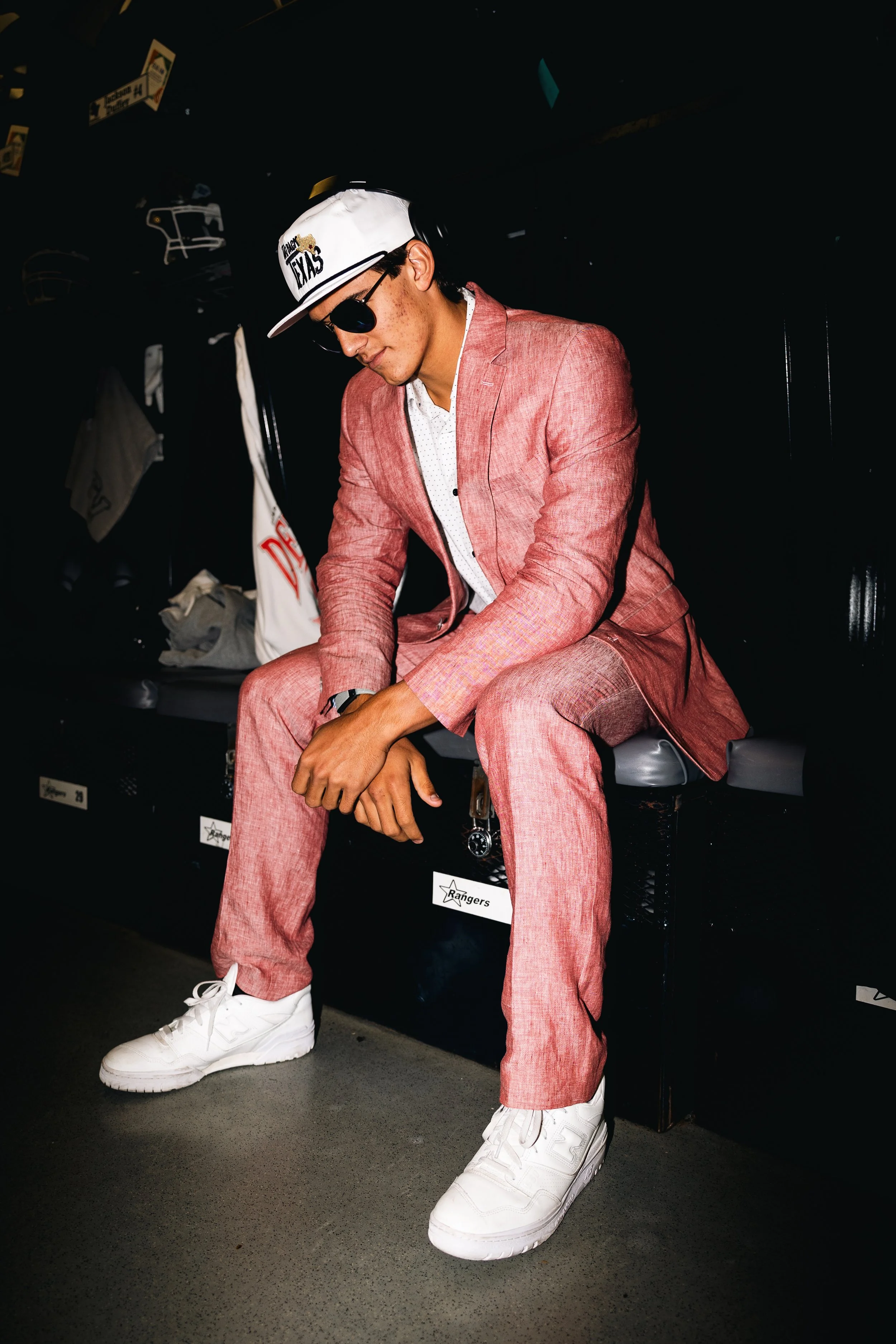 A young man wearing a pink suit, white sneakers, black sunglasses, and a white cap with text, sitting in a dark locker room.