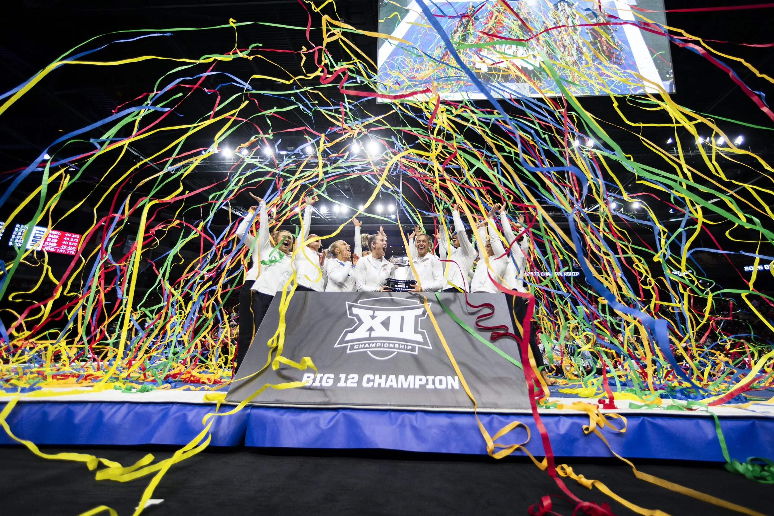The university women's gymnastics team celebrating their victory at the Big 12 Championship surrounded by colorful streamers and confetti in an indoor sports arena.
