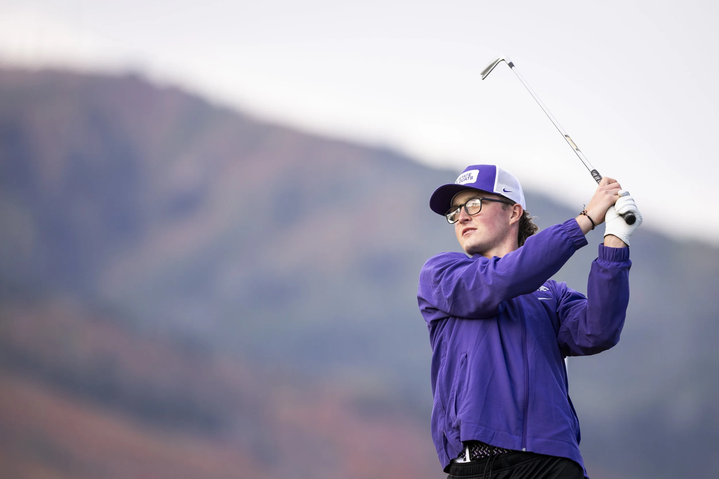 A young male golfer wearing glasses, a purple cap, and a purple jacket swings a golf club on a golf course with mountains in the background.