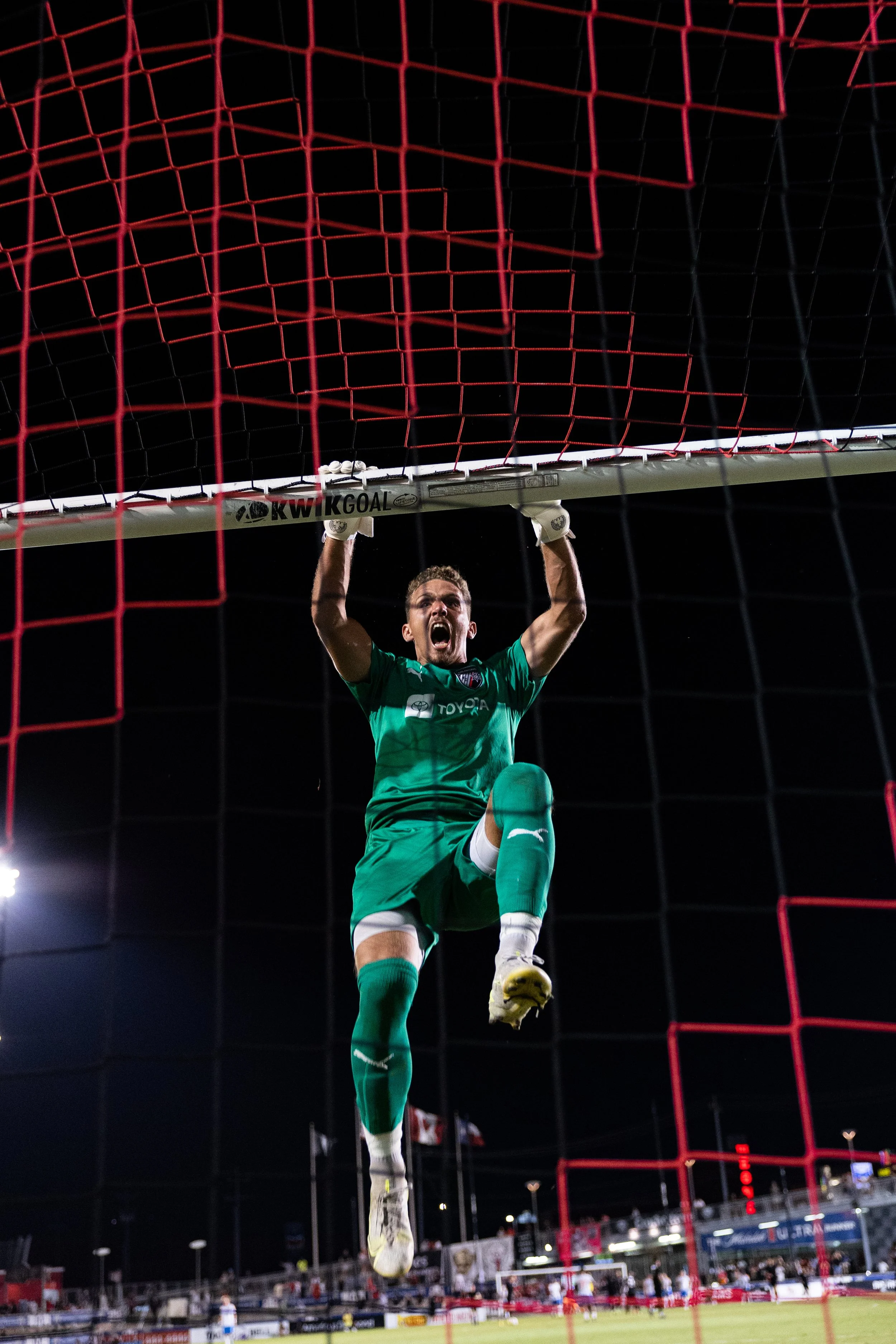 A soccer goalkeeper wearing a green uniform and gloves, hanging from the crossbar of the goal with an intense expression during a night match.