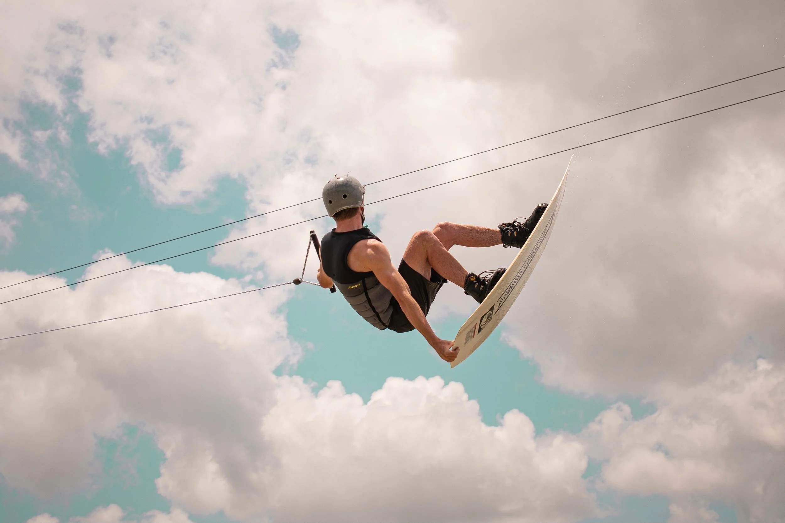 Person wearing a helmet and harness riding a wakeboard on the cable ski over water with cloudy sky background.