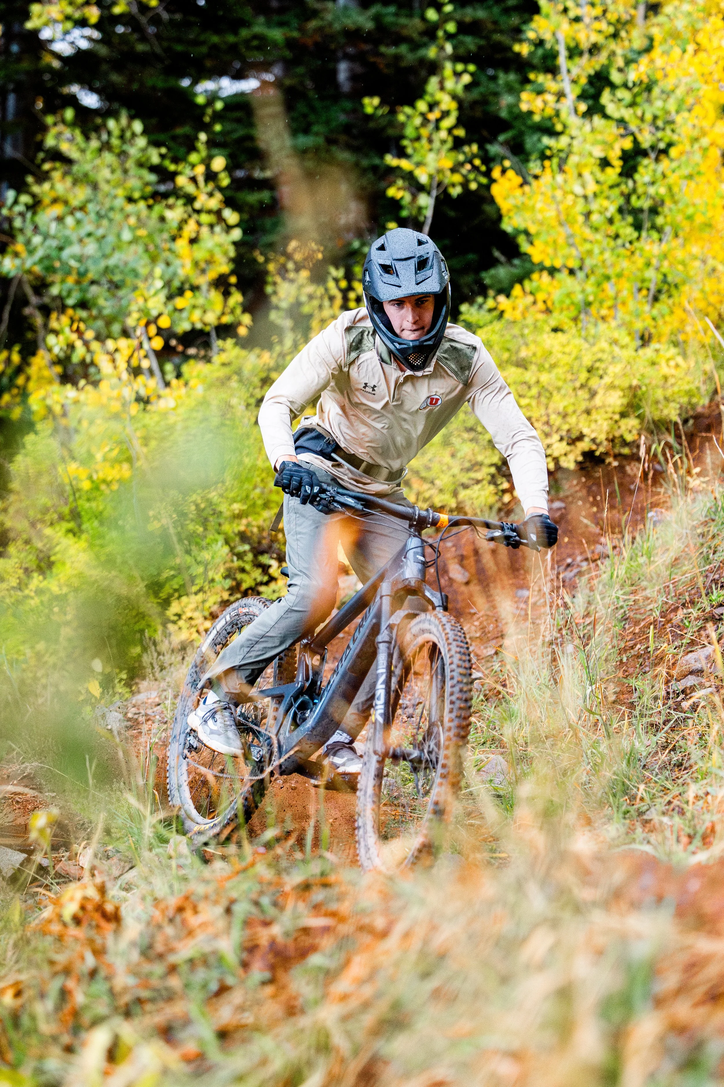 Person mountain biking on a dirt trail surrounded by autumn foliage, wearing a helmet and outdoor biking gear.