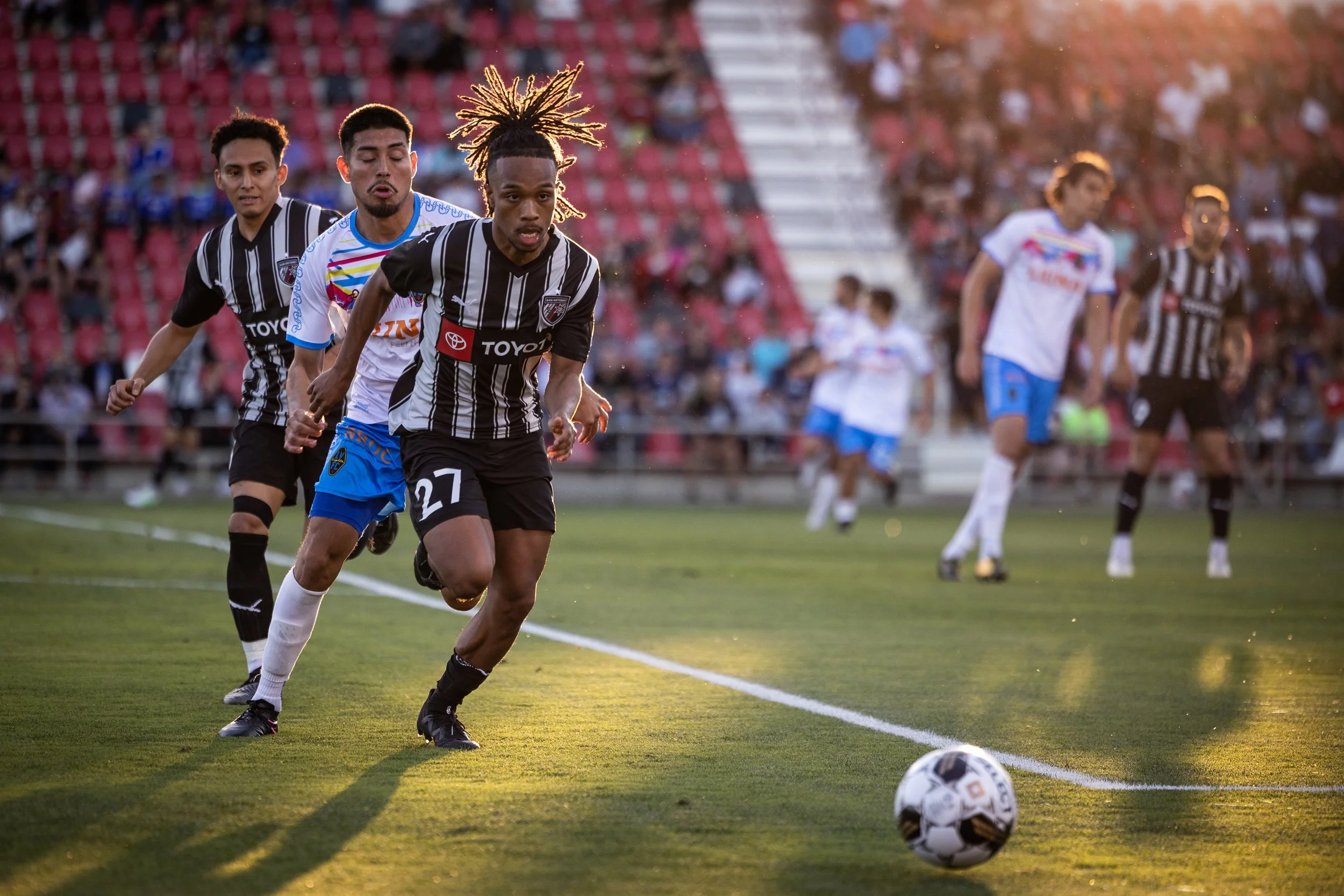 Soccer players running on a field chasing a ball, with spectators in the stands, during a match at sunset.