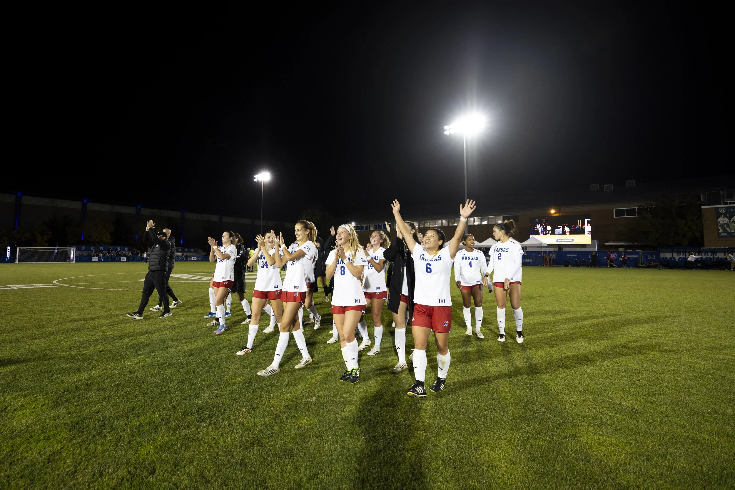 A group of women soccer players celebrating on a field at night under bright stadium lights, wearing white jerseys with blue text and red shorts.