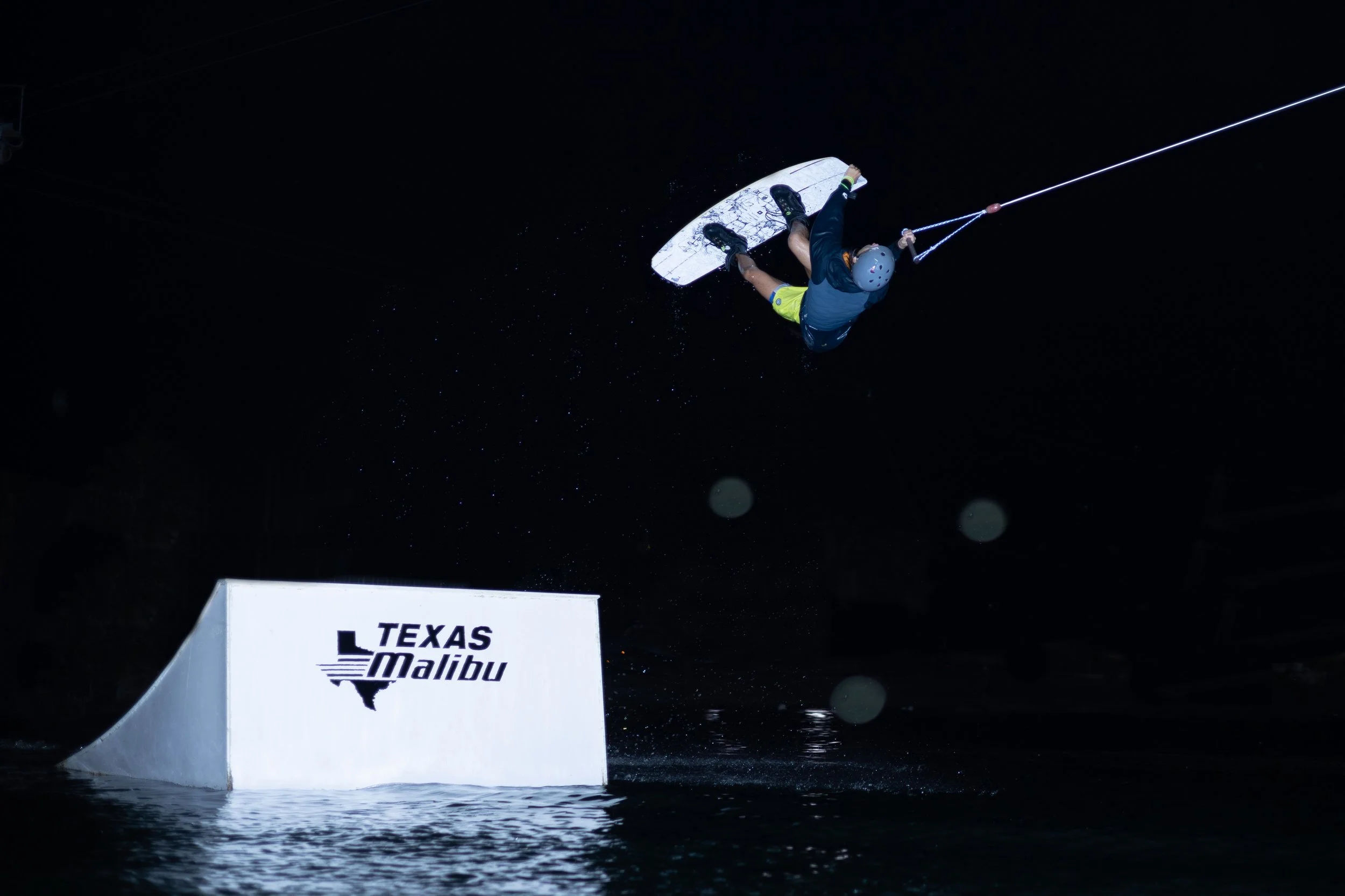 A person wakeboarding at night, performing a jump over a ramp with Texas Malibu logo, water splashing below.