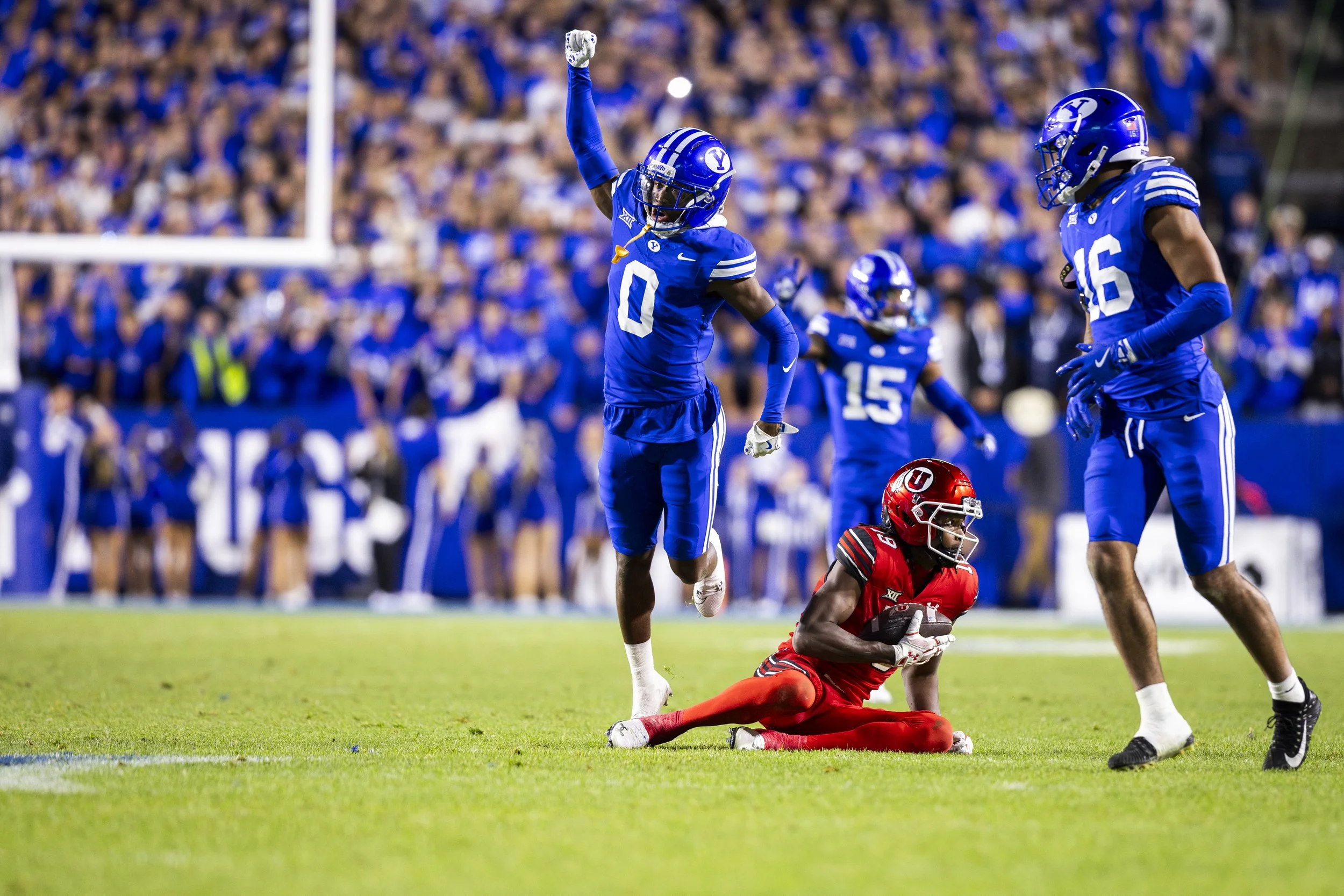 A college football game with players in blue and red jerseys on the field. One player in a red jersey is on the ground holding the football, while two players in blue jerseys are nearby. The crowd is visible in the background.