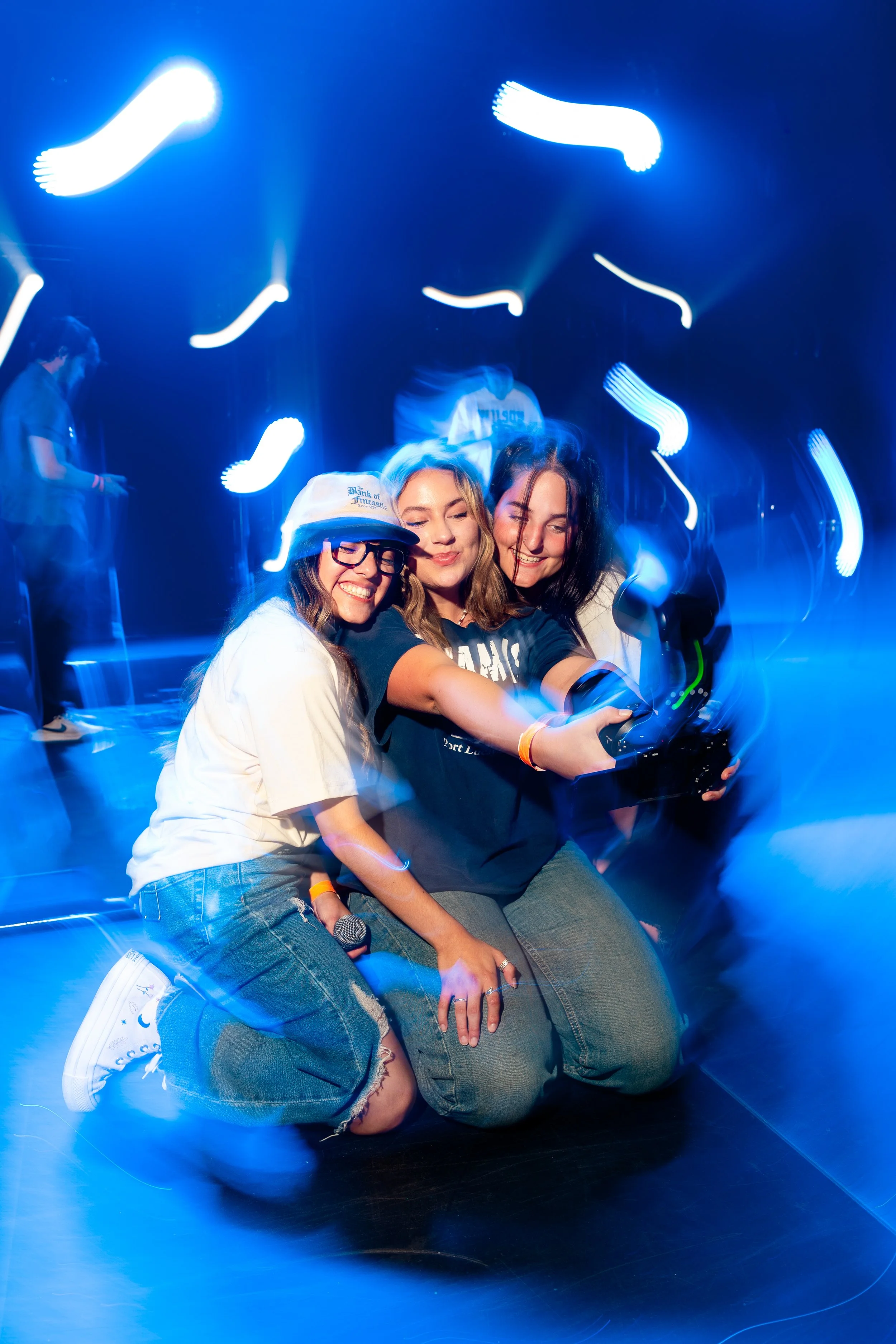 Three young women smiling and taking a selfie together on a stage at a music event, with blue stage lighting and blurred figures in the background.