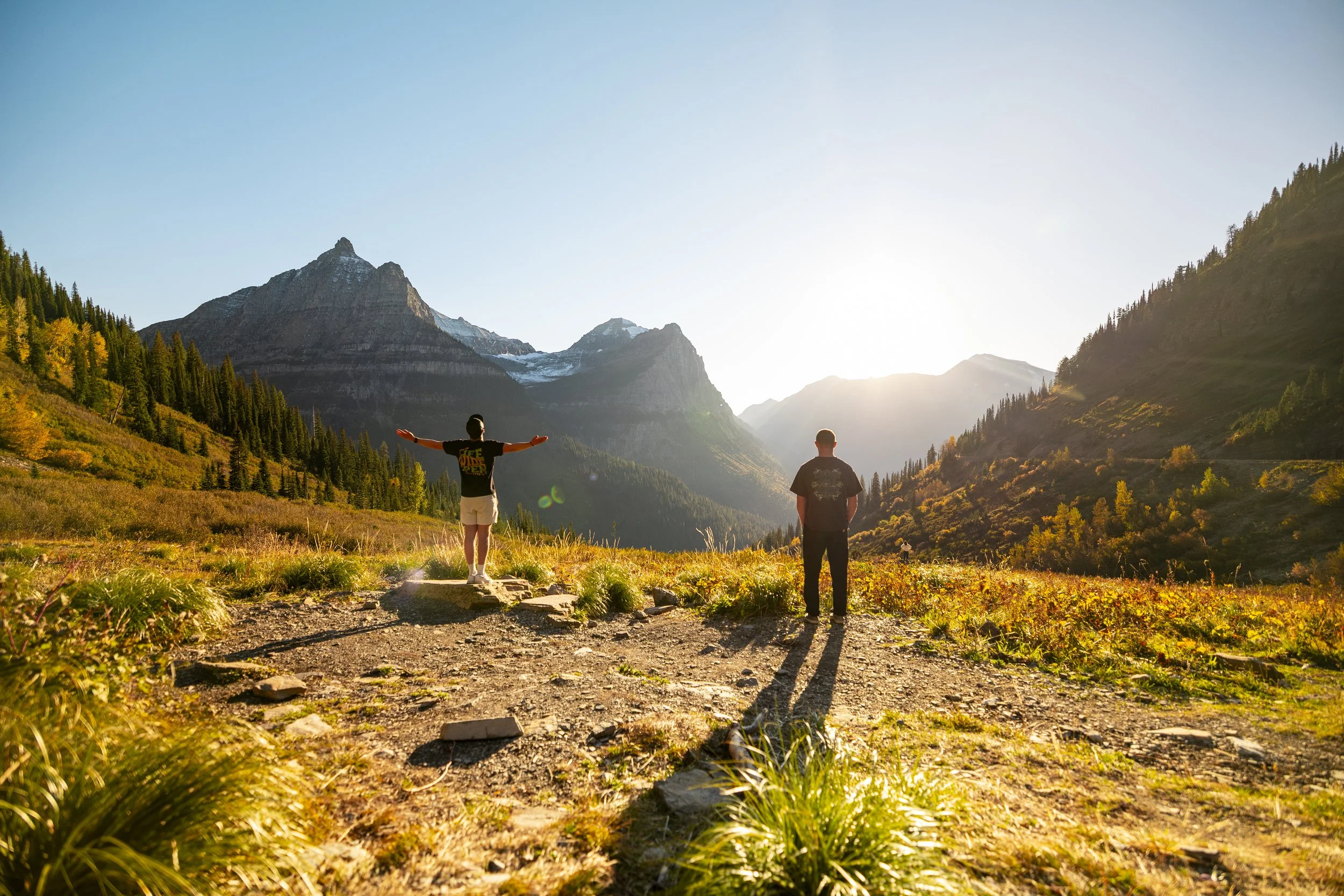 Two people standing on a trail in a mountainous landscape, one with arms outstretched, during sunset with mountains and trees in the background.