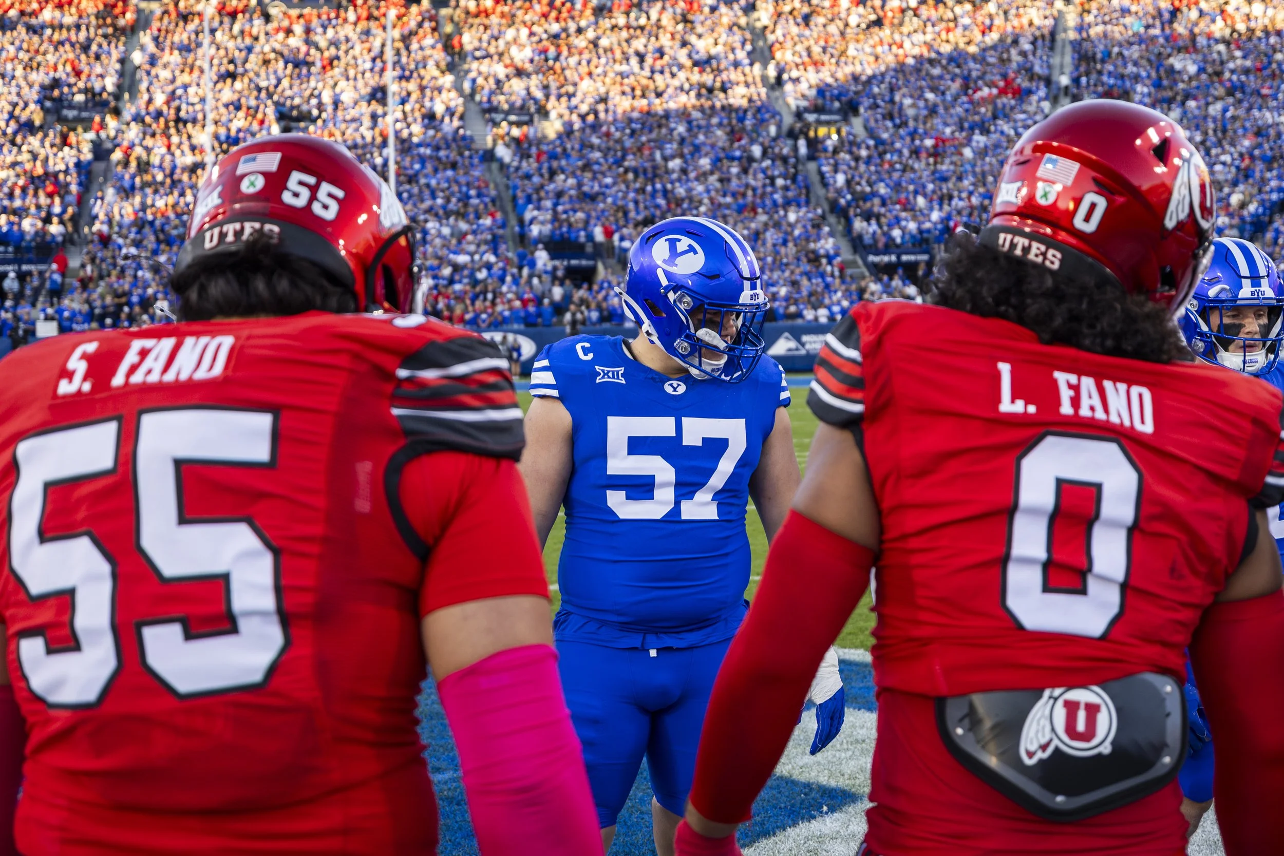 American football players facing each other on the field before a game, with a crowd in the stadium background.