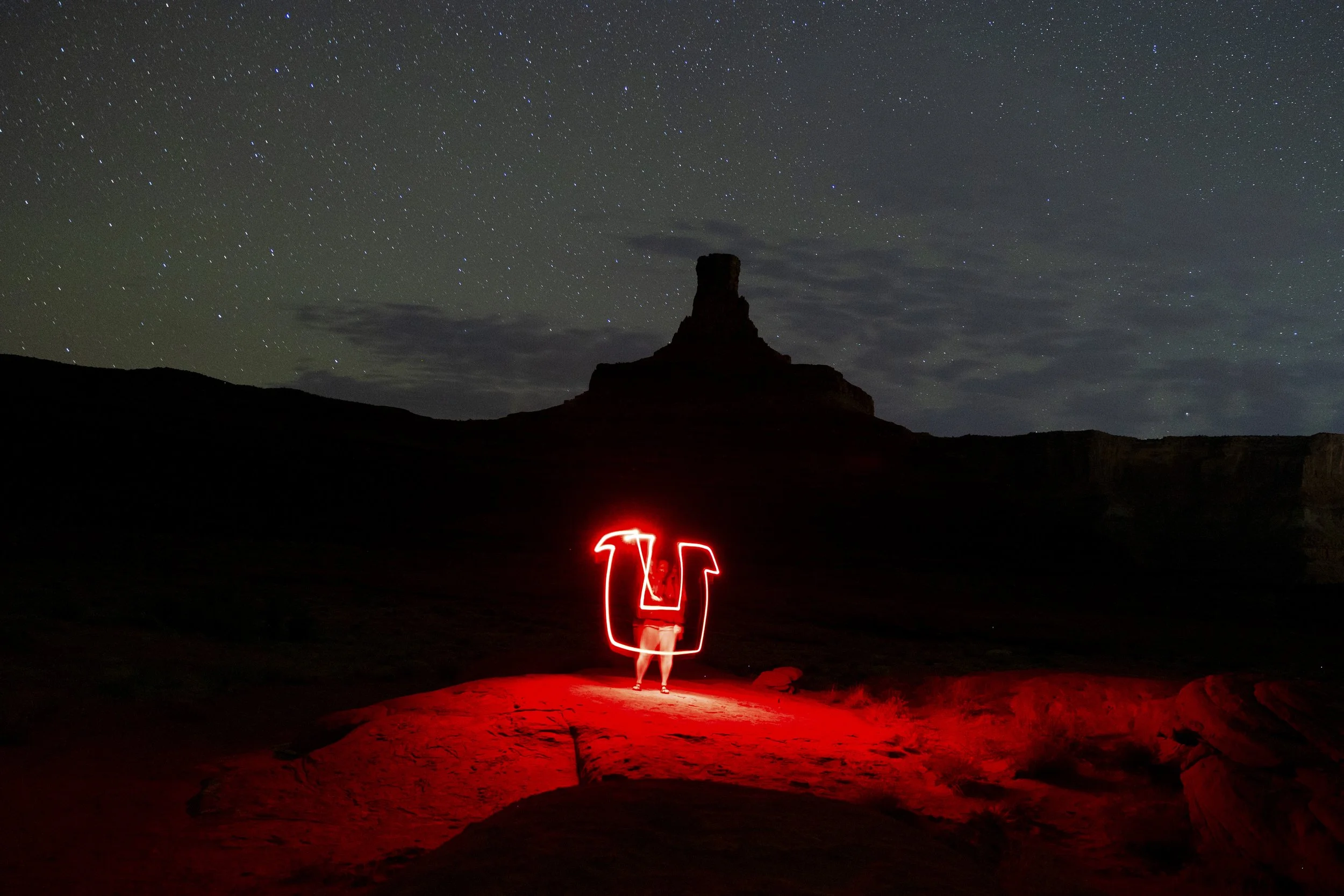 A person standing on a desert landscape at night, illuminated by red light, with a large rock formation in the background under a starry sky.