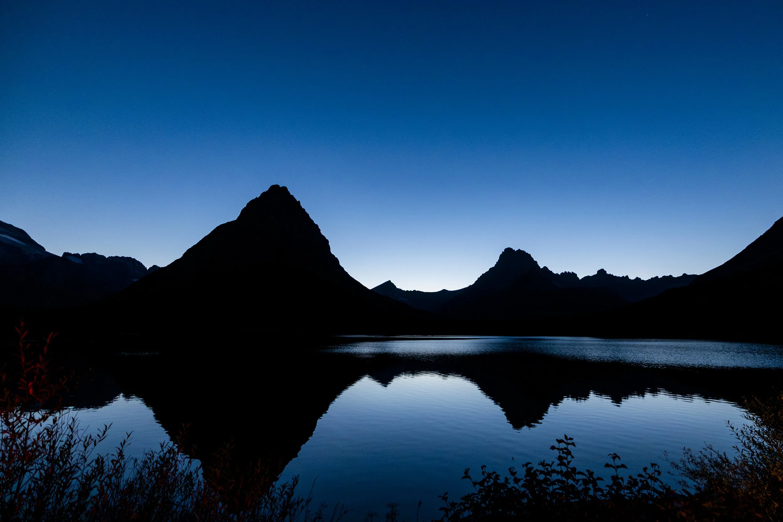 A silhouette of mountain peaks reflected in a calm lake during dusk or dawn.