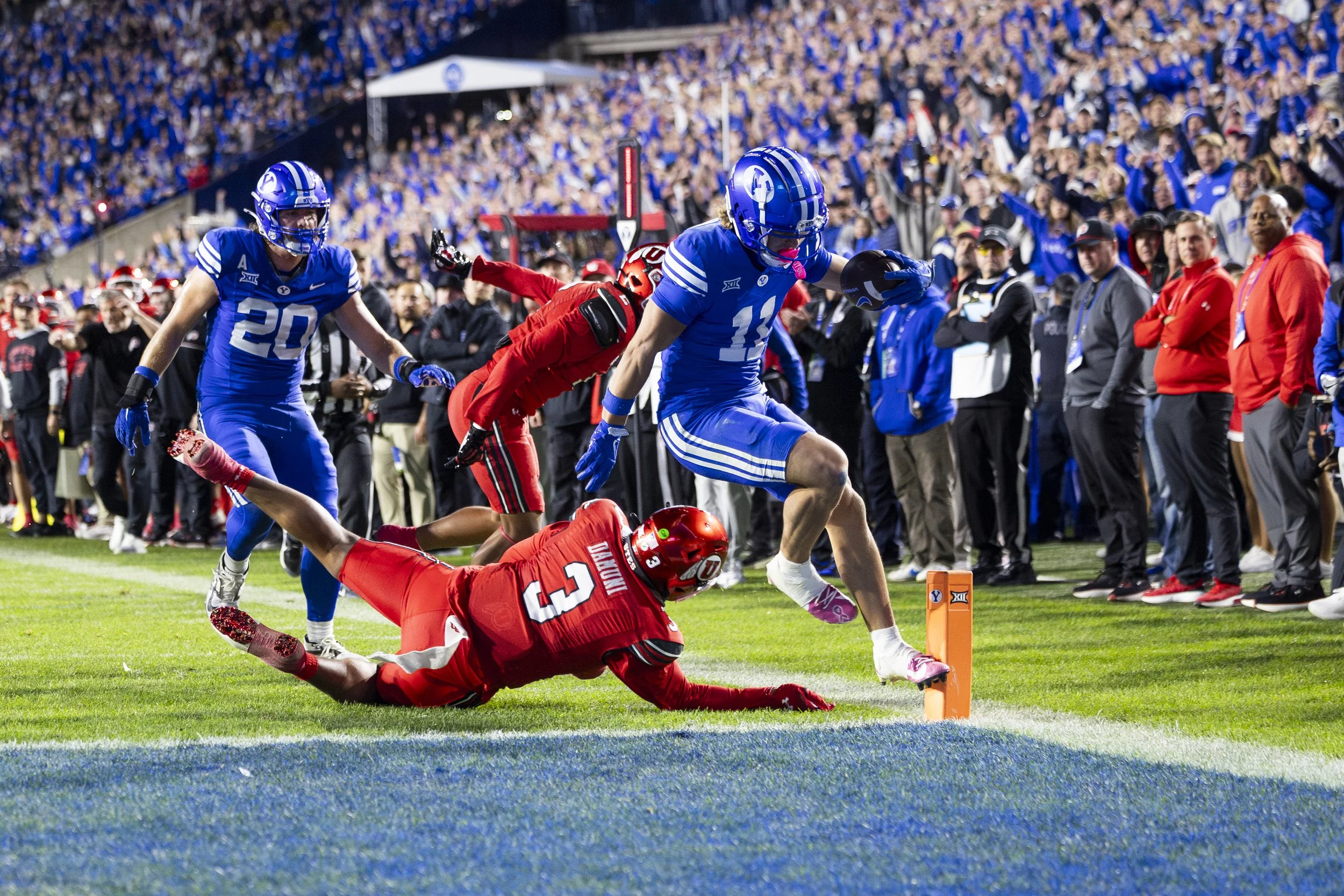 A college football game with players and spectators. A player in a red uniform is diving towards the end zone, while a player in a blue uniform tries to reach the ball first. The crowd in the background watches the play.