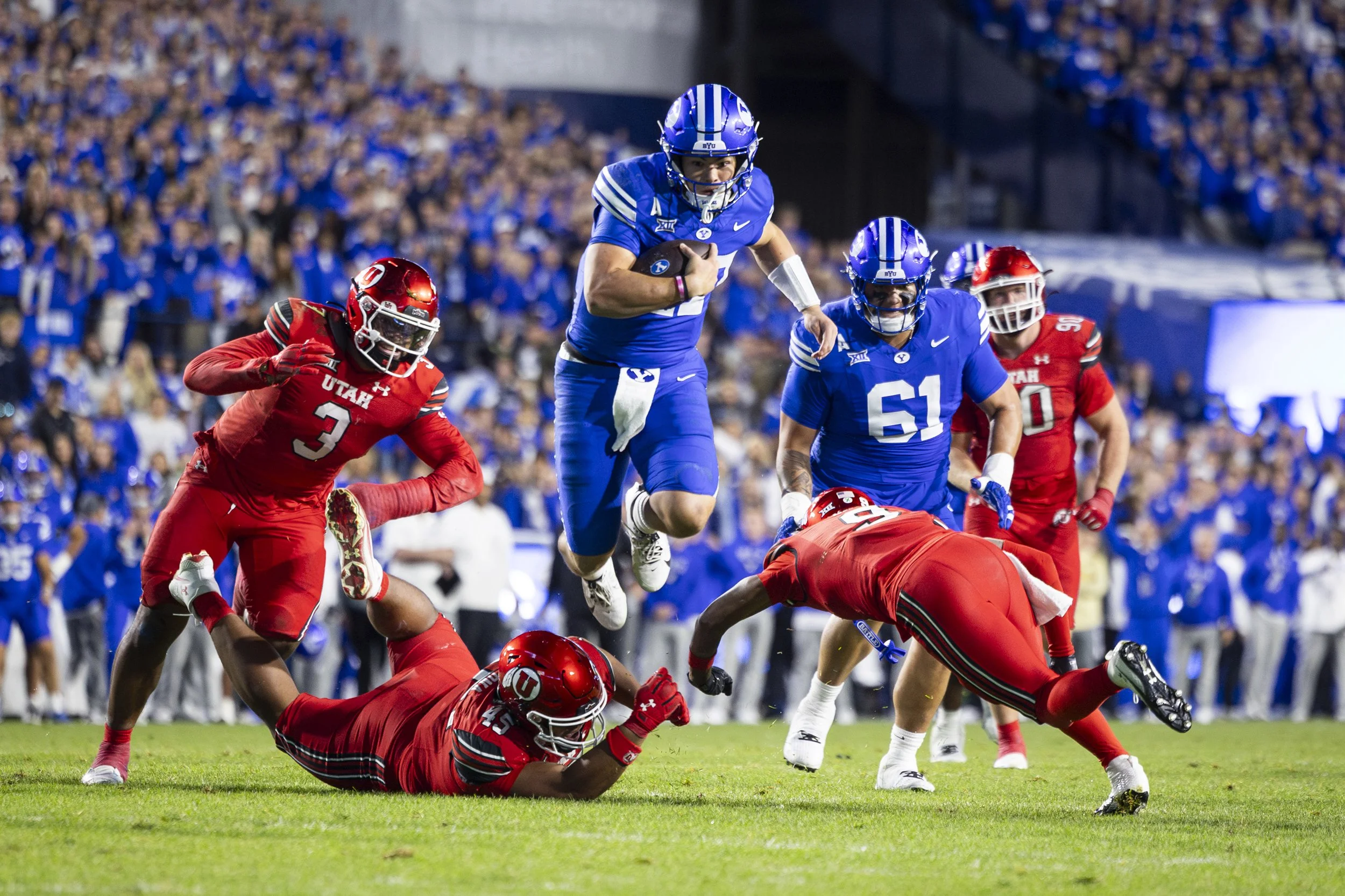 American football game with players in red and blue uniforms on the field, one blue player jumping over a fallen red player, with other players and crowd in the background.