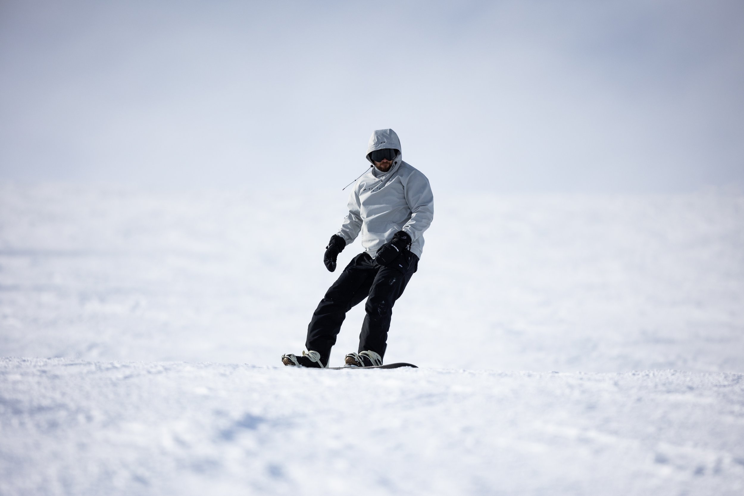 Person snowboarding on a snowy slope, wearing a white hooded jacket, black pants, gloves, sunglasses, and a hood, with cloudy sky in the background.