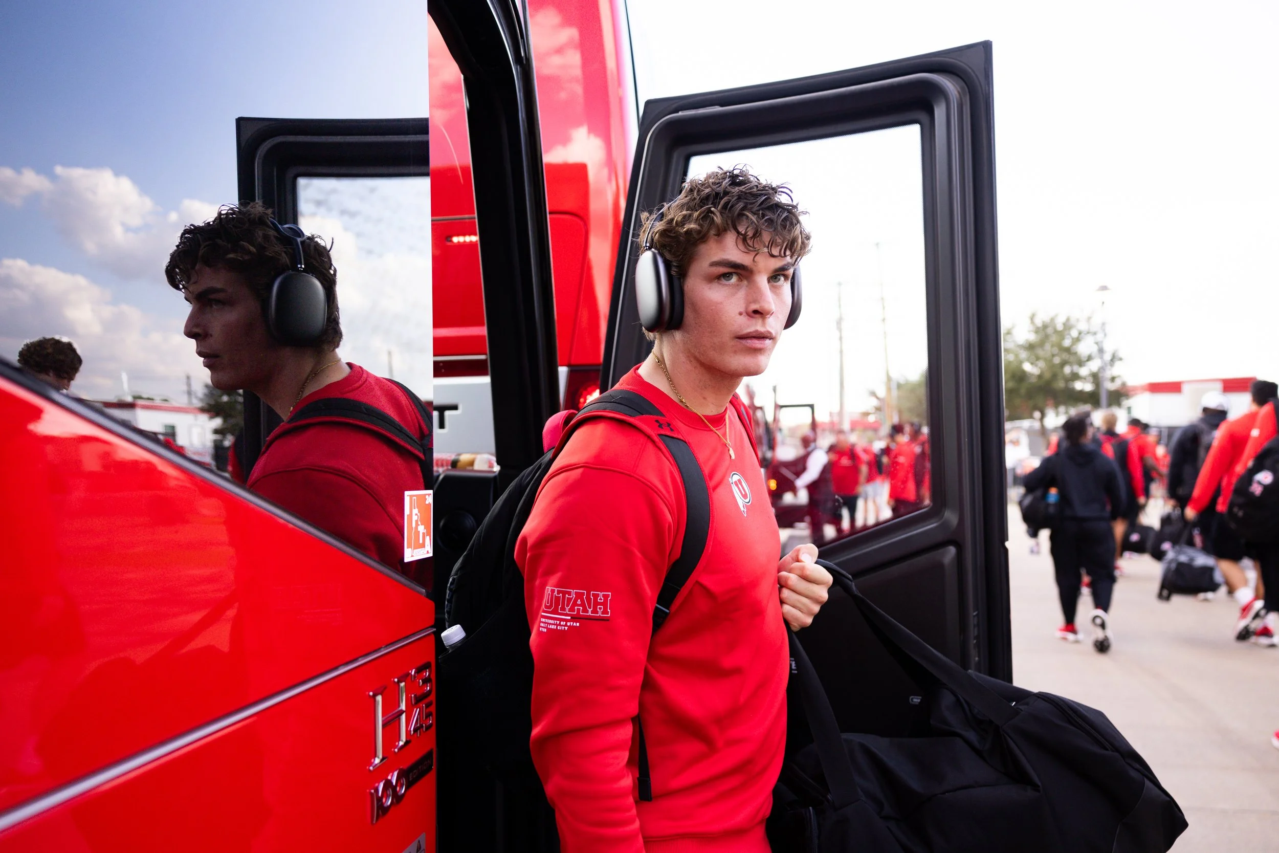 Young man with curly hair wearing headphones, a red sports hoodie with 'UTAH' and 'Utes' on the sleeve, carrying a black duffel bag, standing near a red fire truck at a sports team event.