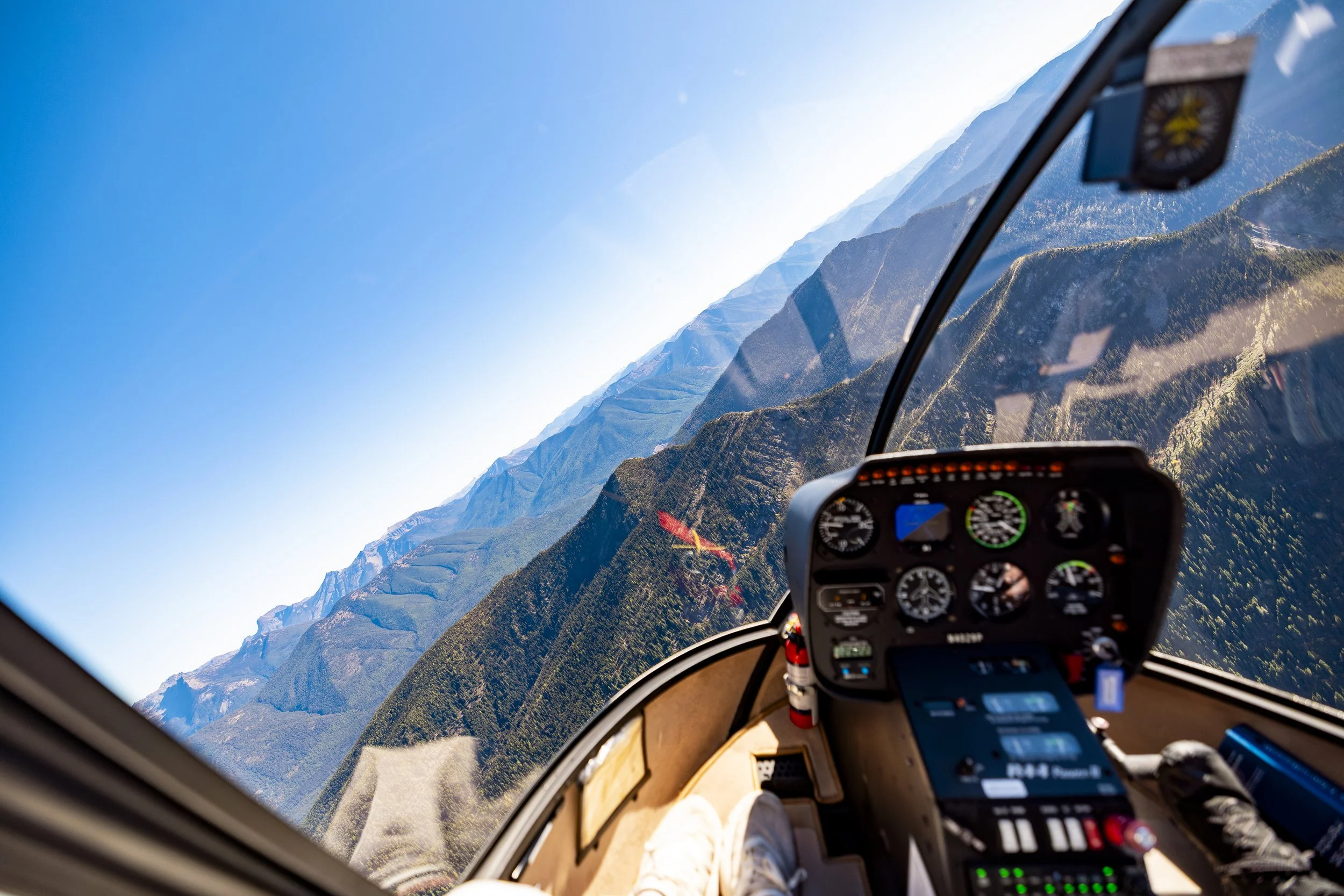Inside a helicopter cockpit flying over a mountainous landscape with clear blue sky, showing various instruments and dashboard controls.
