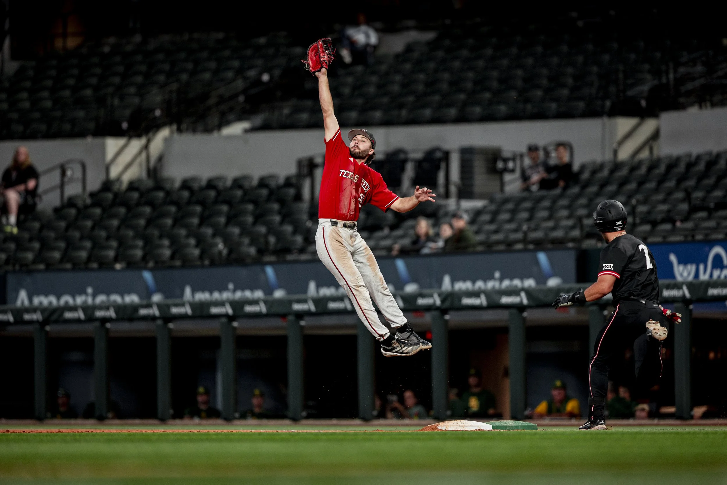 A baseball player in a red Texas Tech uniform makes a leaping catch near second base while a baseman in black and a few spectators sit in the background.
