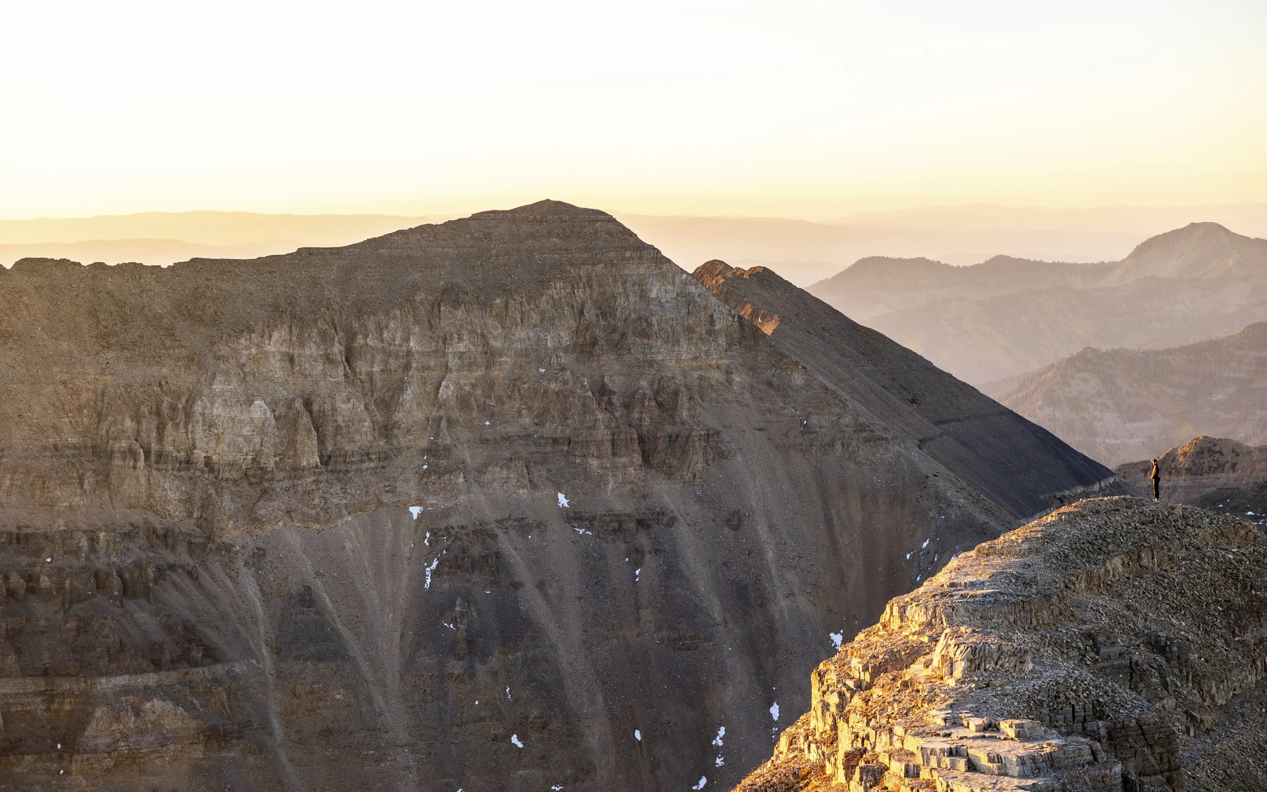 A person standing on a rocky mountain ridge during sunset with a view of layered mountains and valleys in the background.