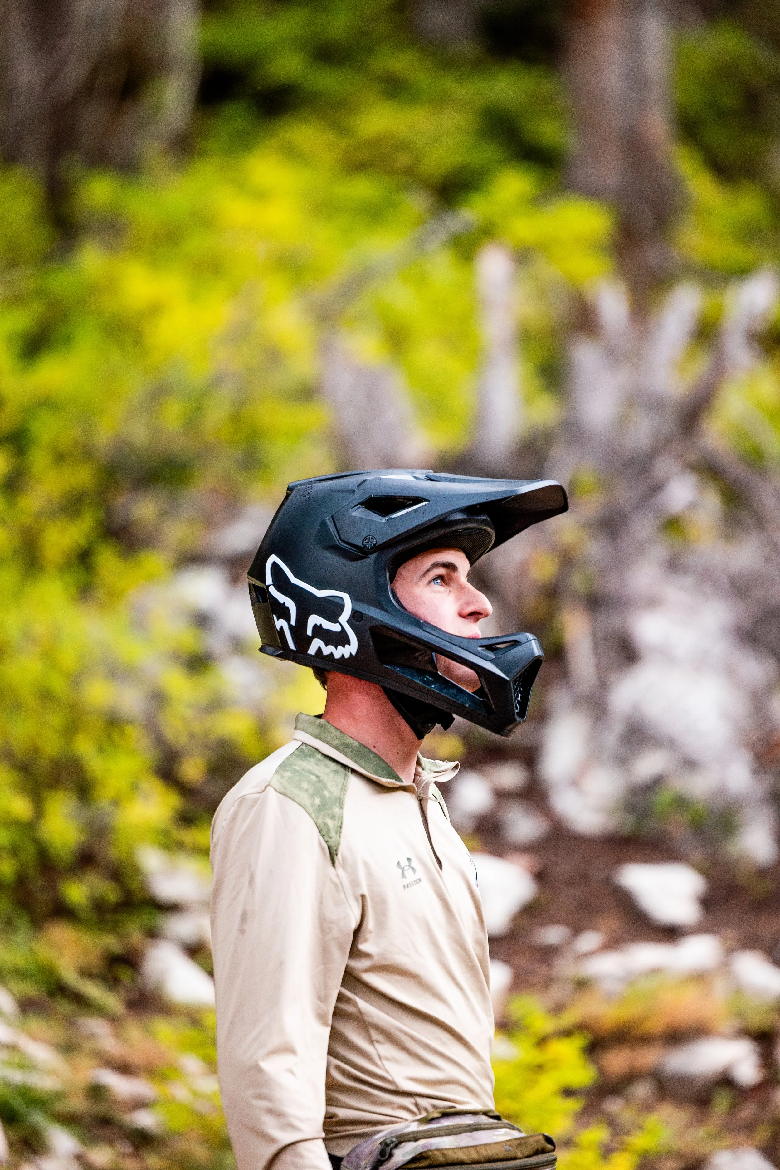 A man wearing a black mountain biking helmet with a white fox logo, standing outdoors with a blurred green and brown forest background, looking to the right.