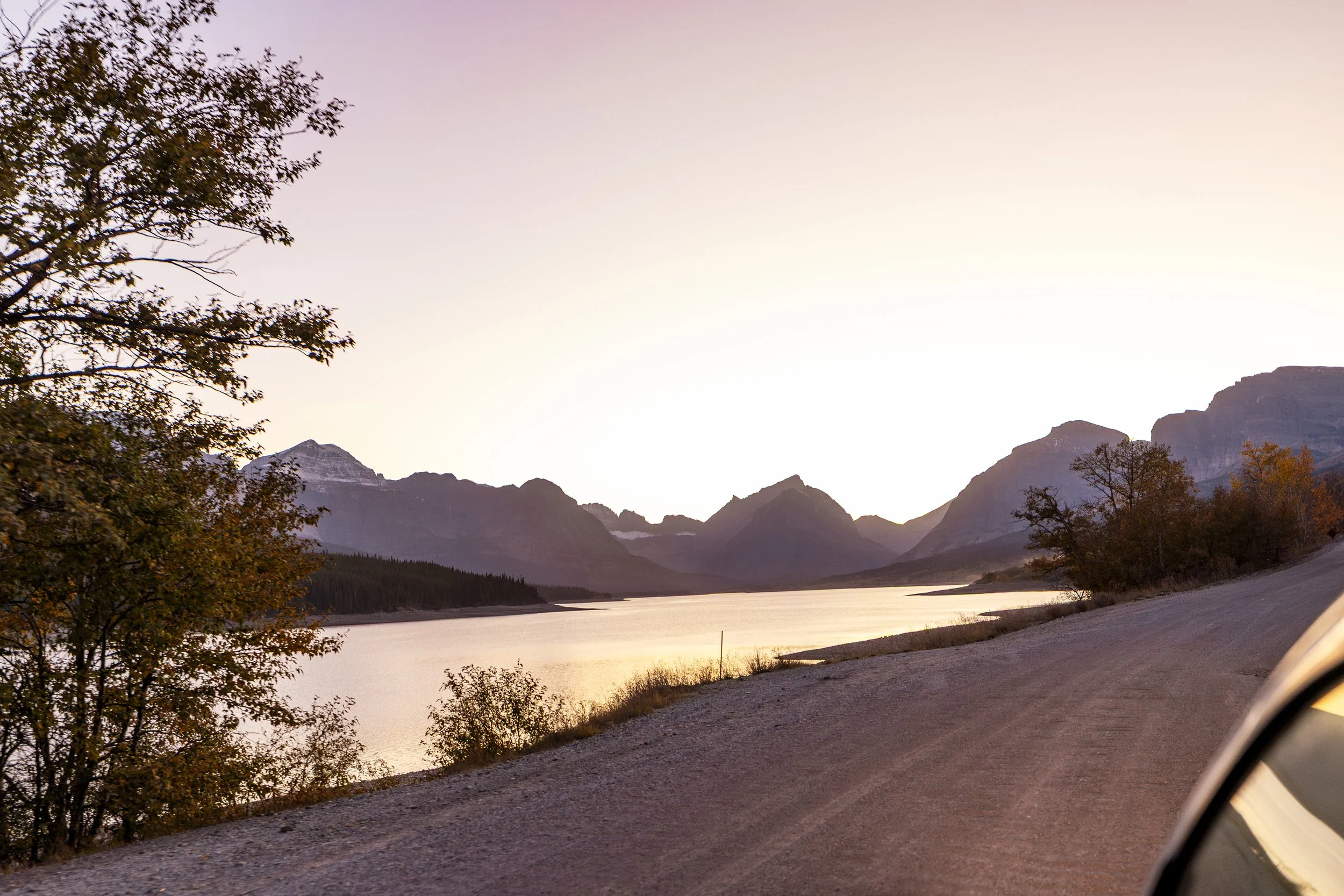Scenic view of a lake with mountains in the distance during sunset, with trees along the shoreline and a dirt road in the foreground.