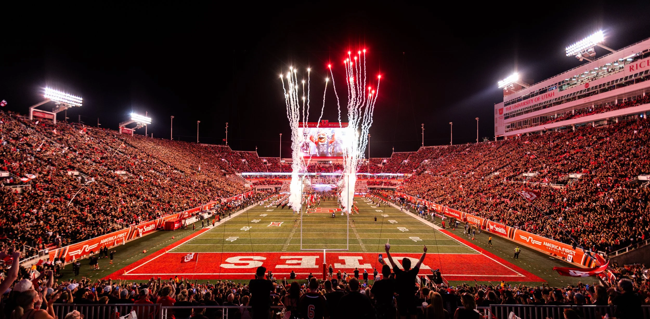Nighttime college football stadium packed with spectators, fireworks erupting in the sky, with players and cheerleaders on the field, and bright stadium lights illuminating the scene.