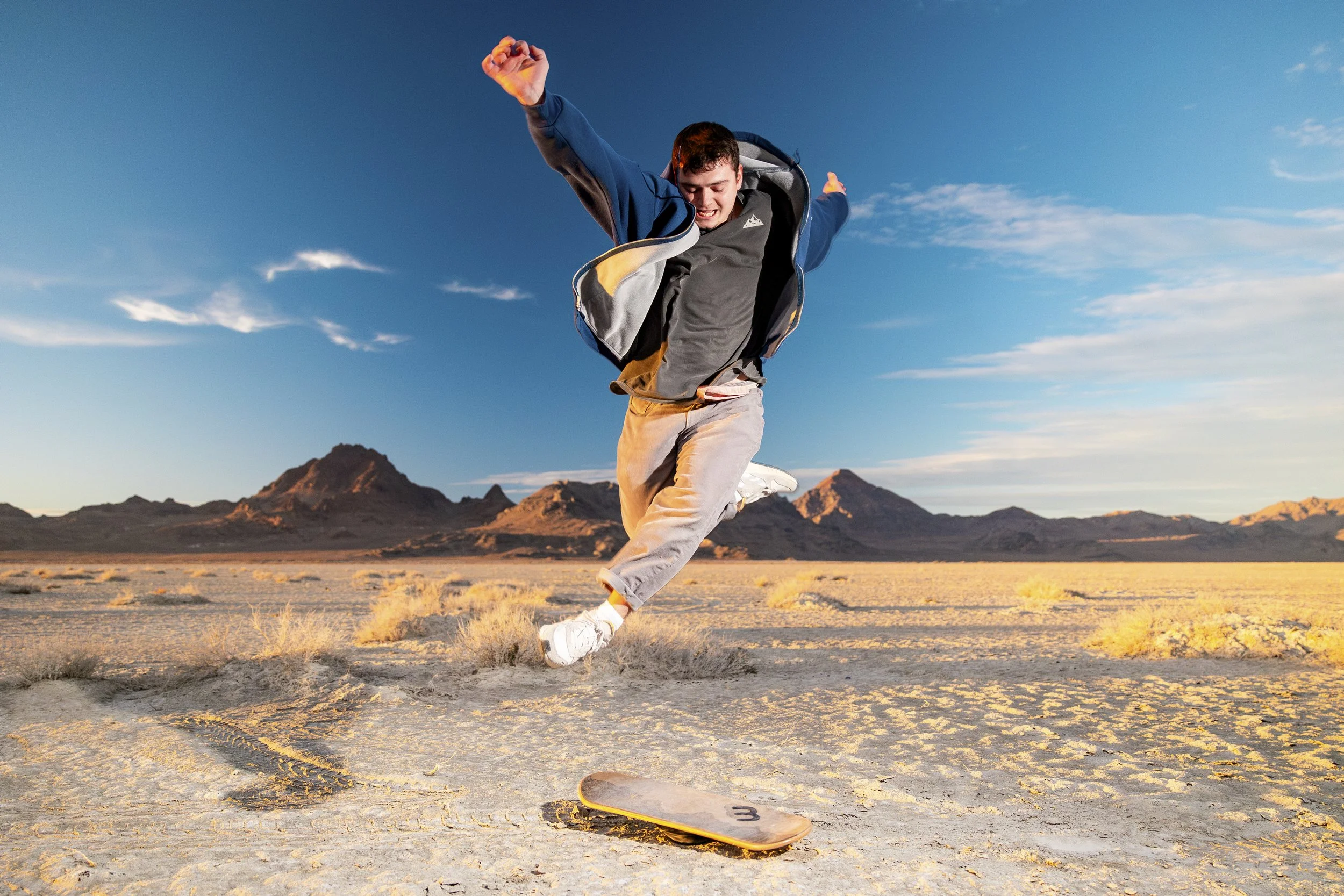 Young man jumping on skateboard in a desert landscape with mountains in the background under a blue sky.