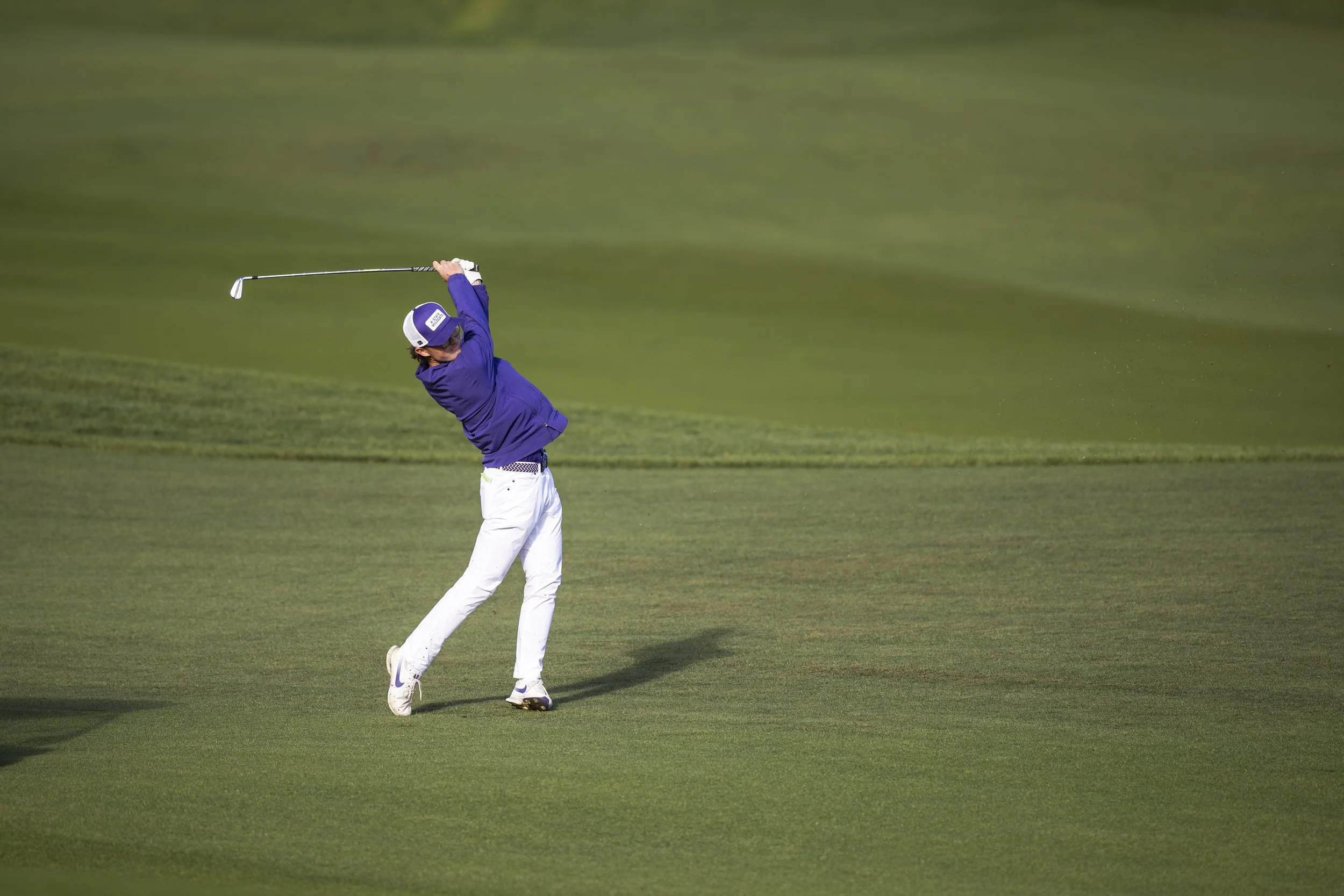 A golfer in a purple shirt and white pants swinging a golf club on a golf course.
