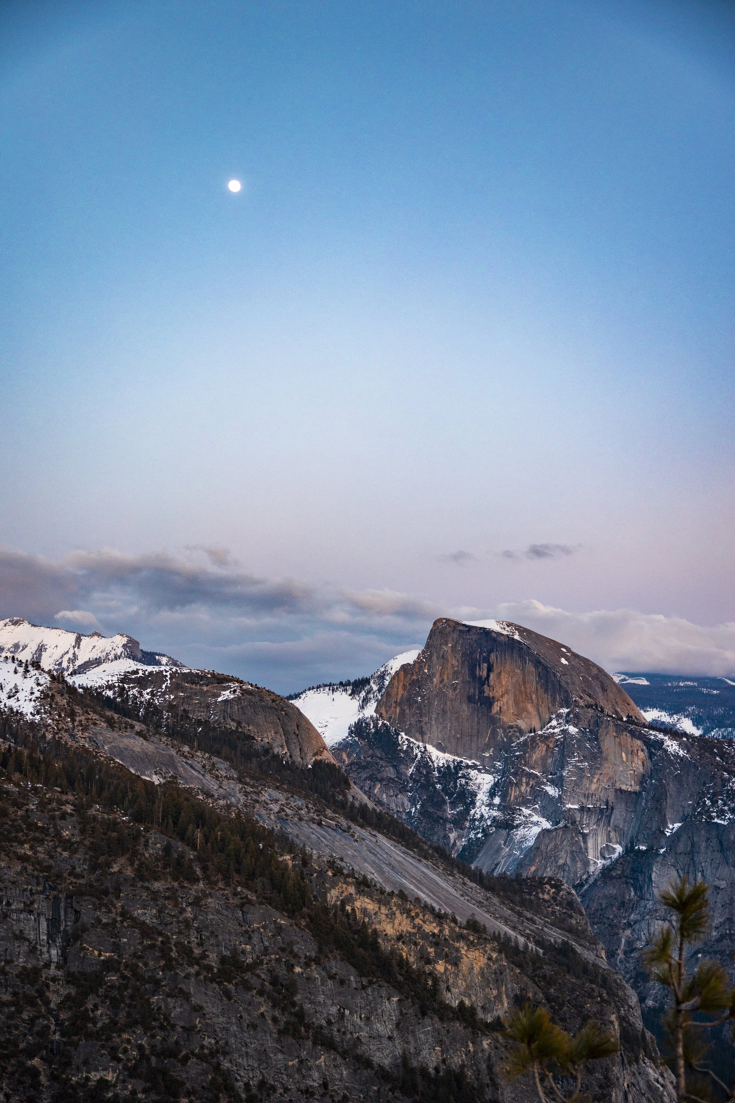 Mountain landscape with snow-capped peaks, a large granite dome, and a rising moon in a twilight sky.