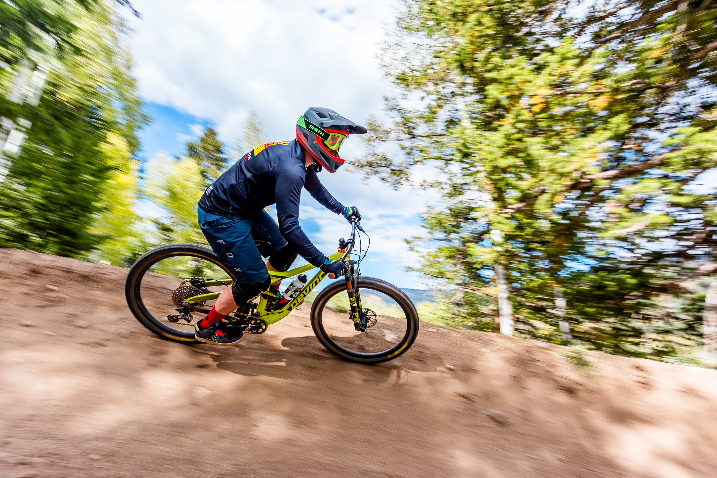 A person mountain biking on a dirt trail surrounded by green trees during daytime, wearing a helmet, goggles, and athletic clothing.