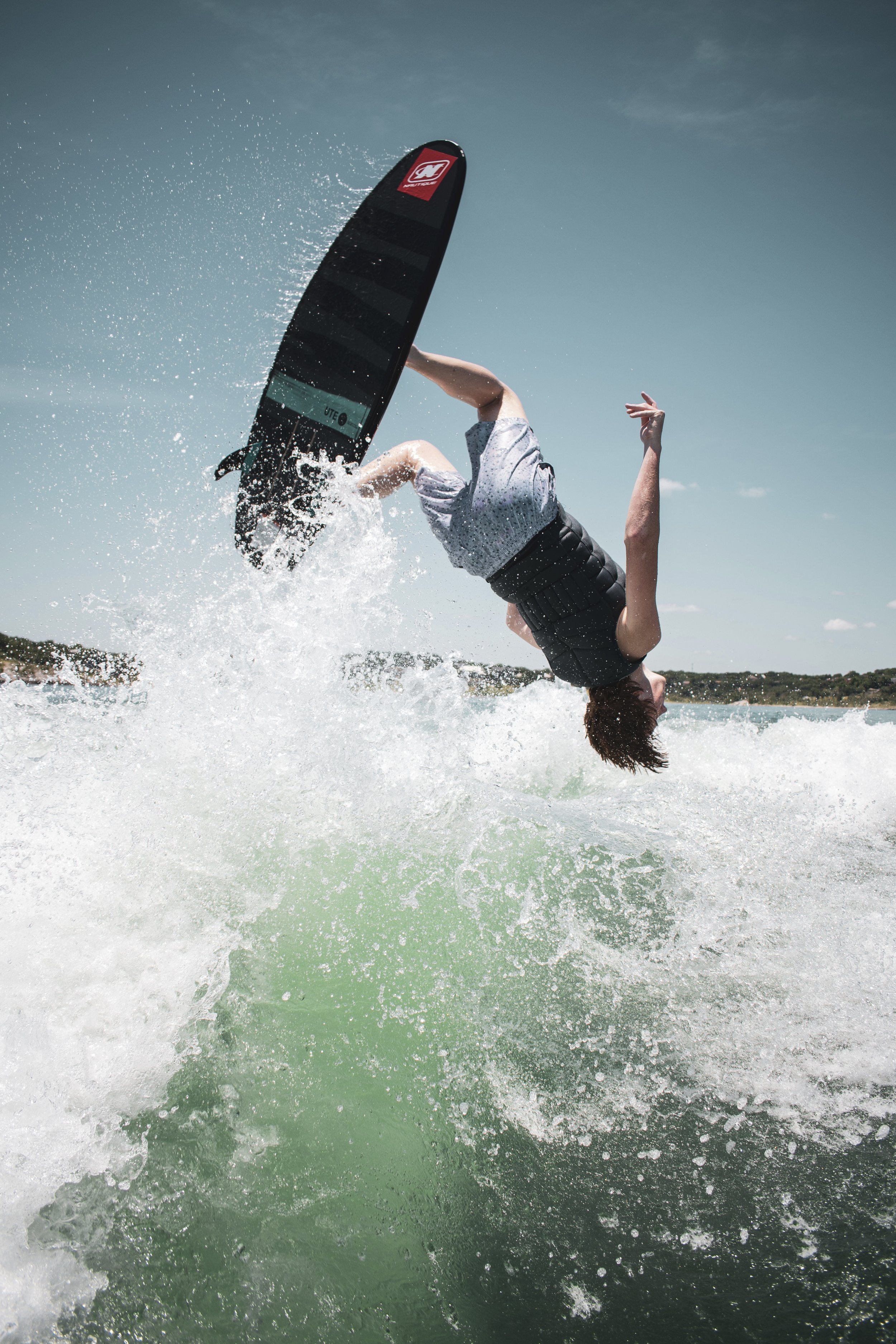 Person in mid-air falling backwards while wakeboarding on water, with splashes and a clear sky in the background.