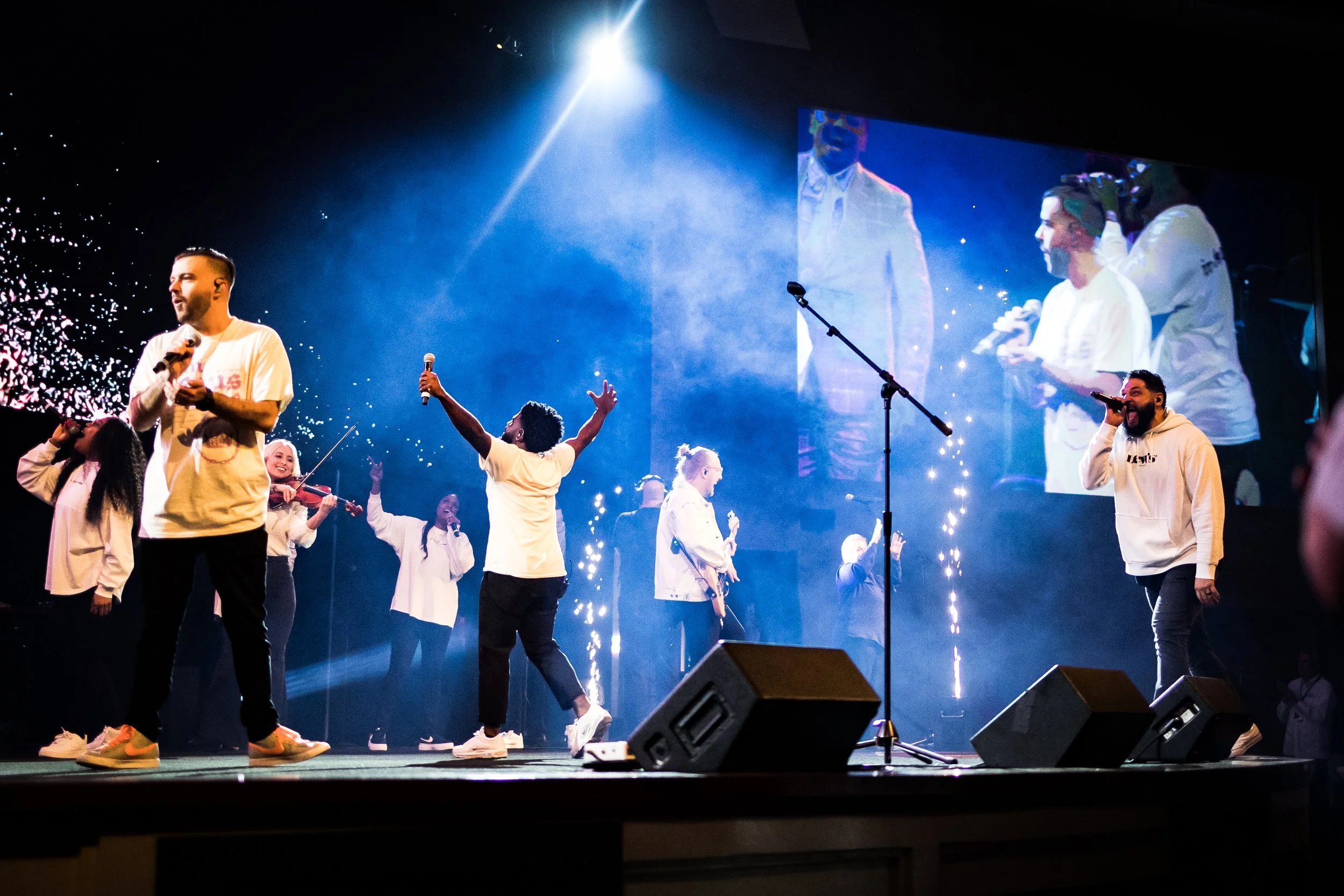 Group of diverse performers on stage during concert, some singing, others playing violin, with large screen behind showing close-ups, purple and blue lighting, and sparklers.