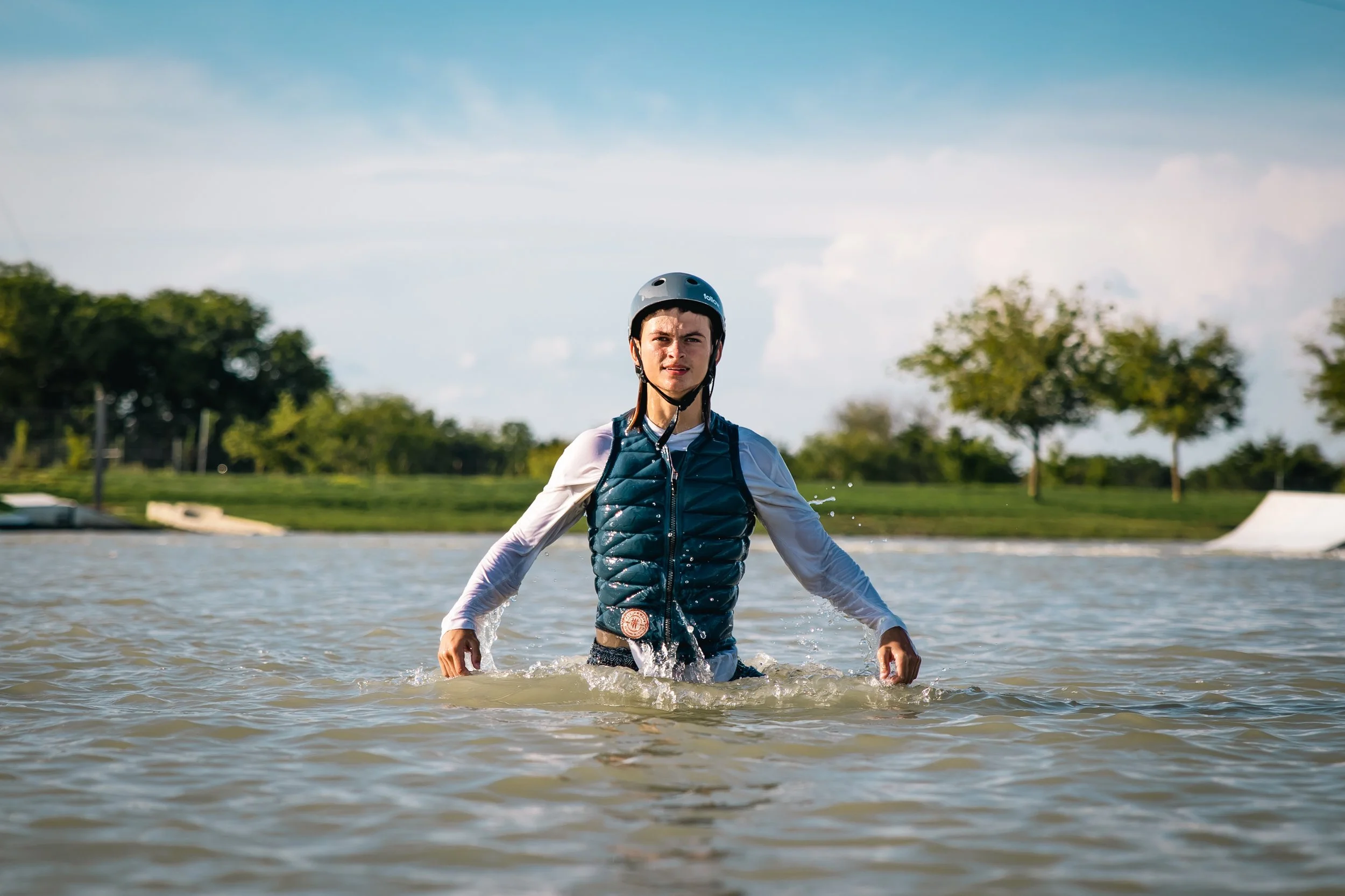 A person wearing a helmet and vest stands waist-deep in water at a park, with trees and a skate ramp visible in the background.