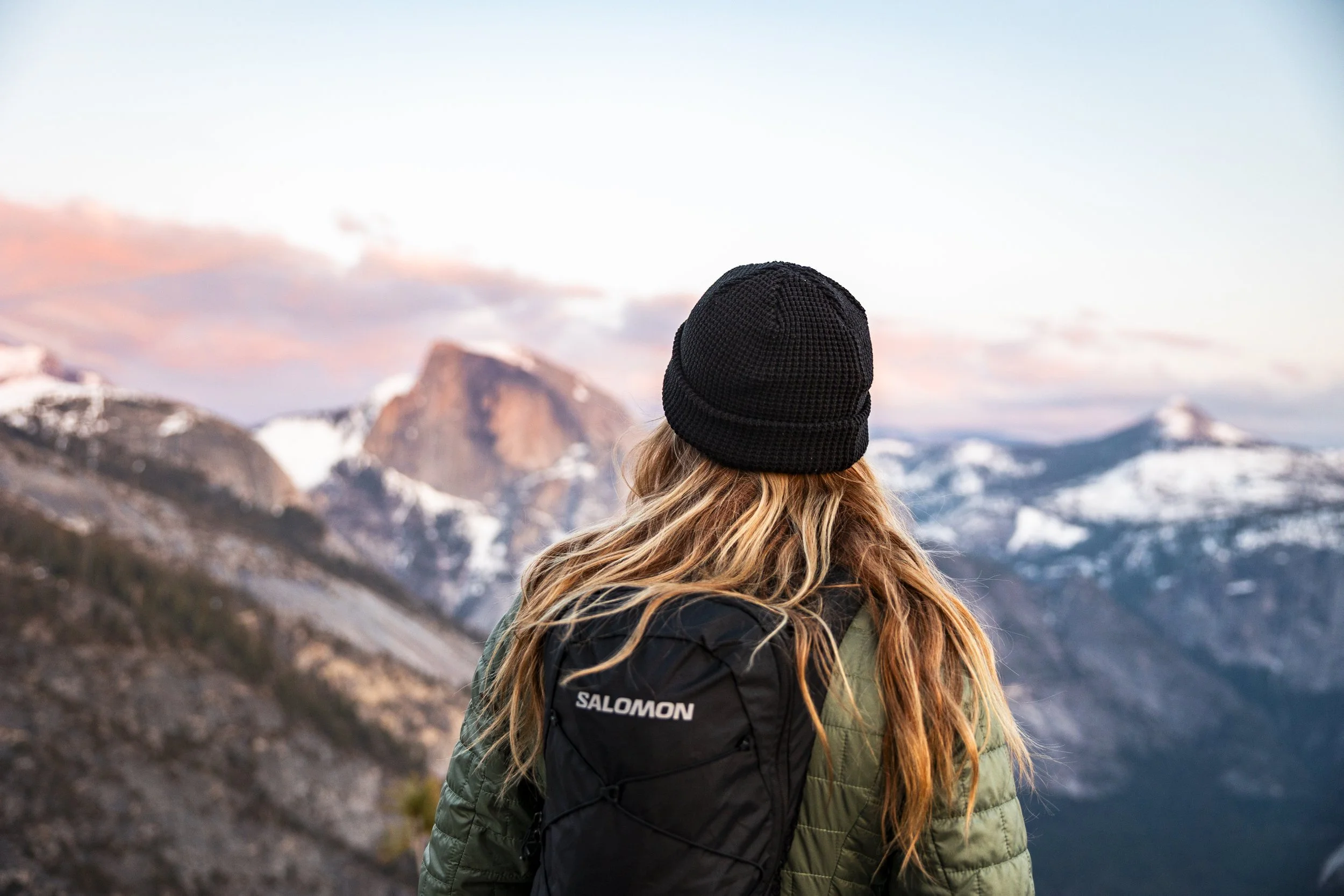 Person with long hair wearing a black knit hat and green jacket, carrying a black Salomon backpack, looking at snow-capped mountains during sunset or sunrise.