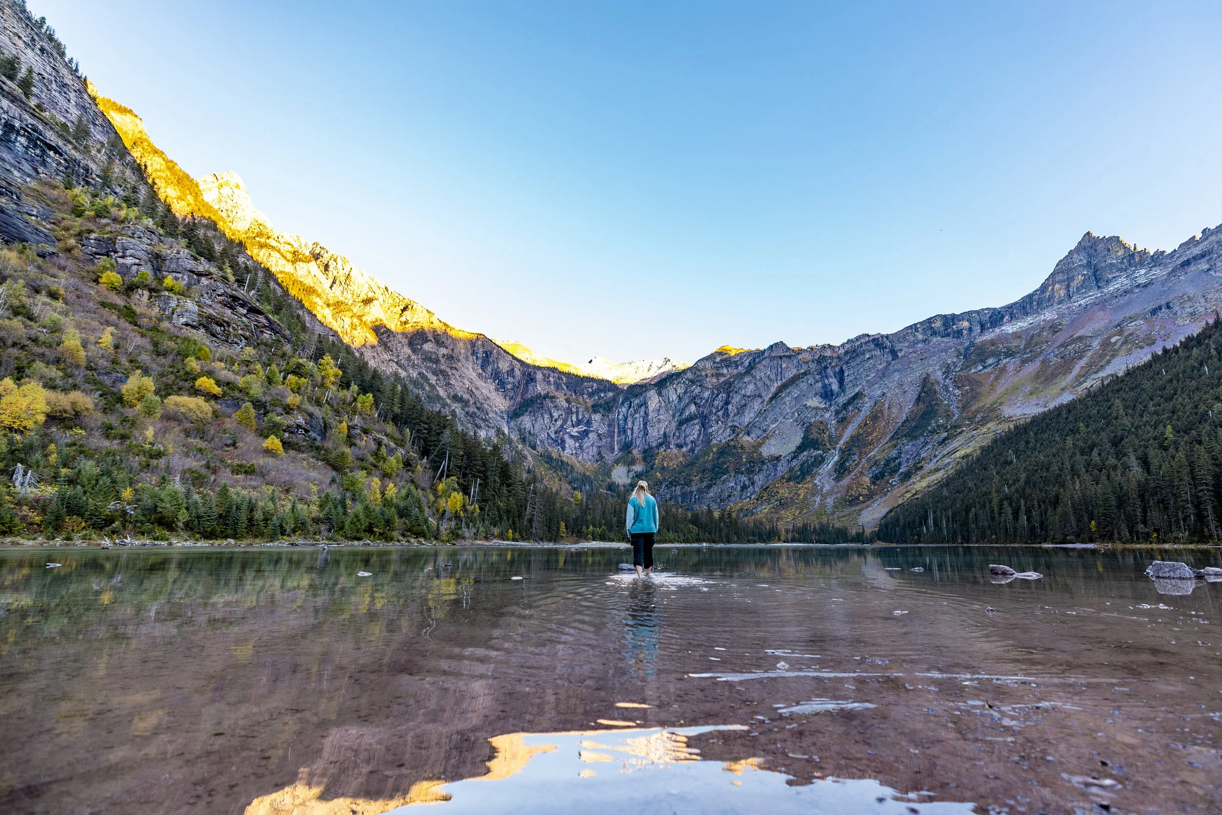 A person standing in a shallow mountain lake surrounded by forested hills and towering mountains under a clear blue sky.