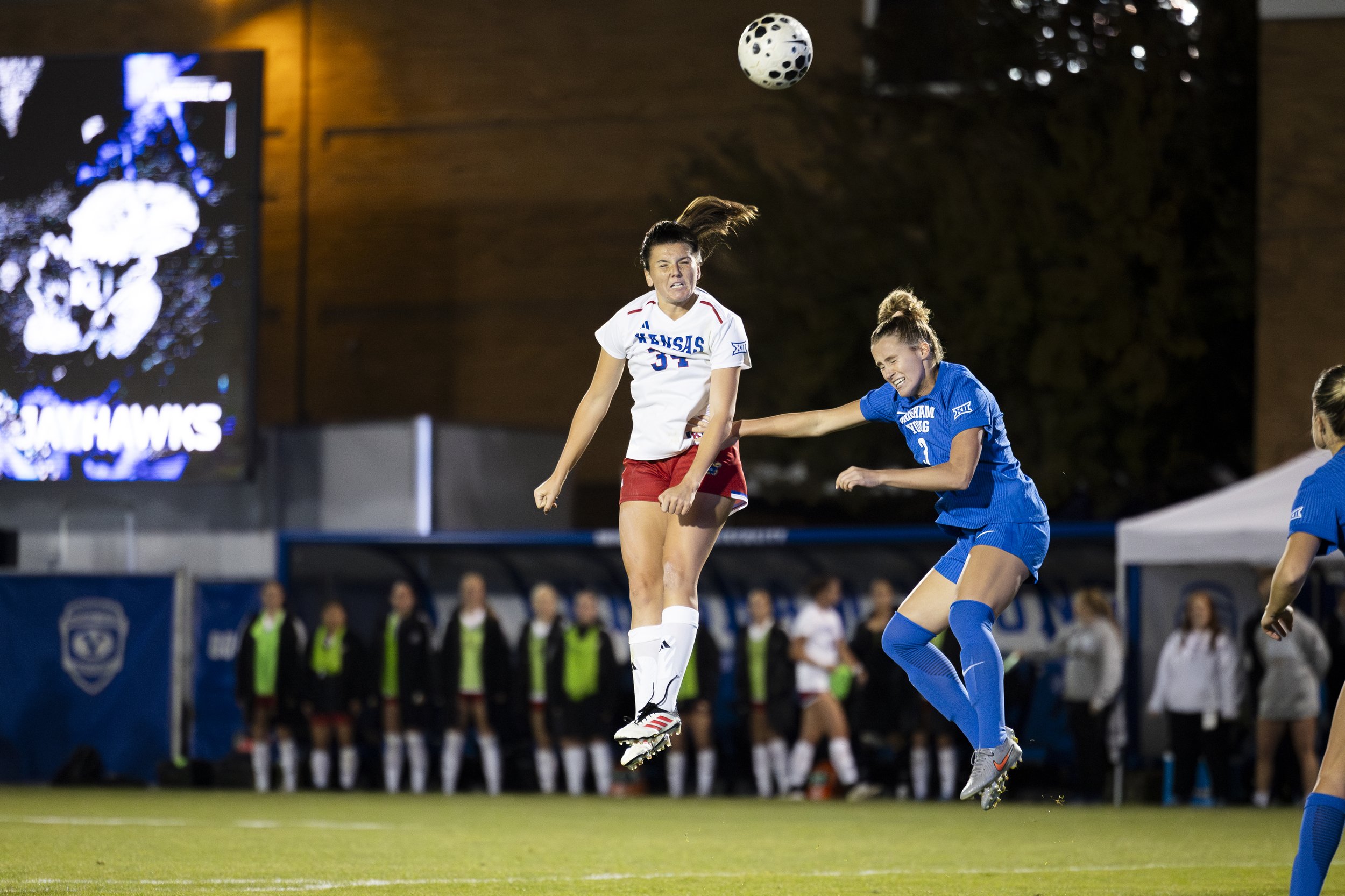 Two female soccer players from opposing teams jump to head the ball during a match. The player in white and red is from Kansas, and the player in blue is from Wichita State. Spectators and team onlookers are visible in the background.