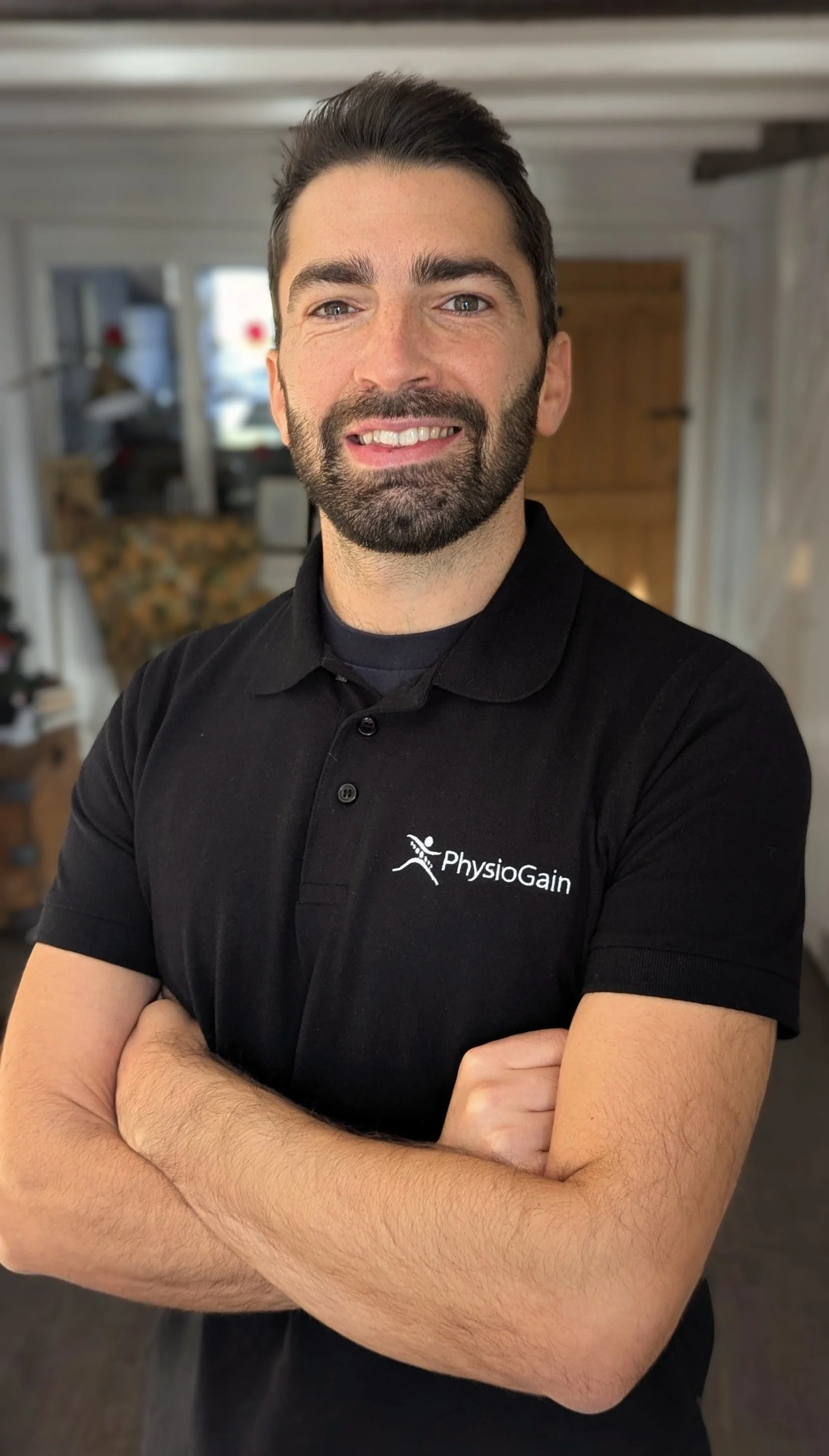 A man with dark hair and a beard, smiling with arms crossed, wearing a black polo shirt with a 'PhysioGain' logo, standing in a cozy indoor setting.