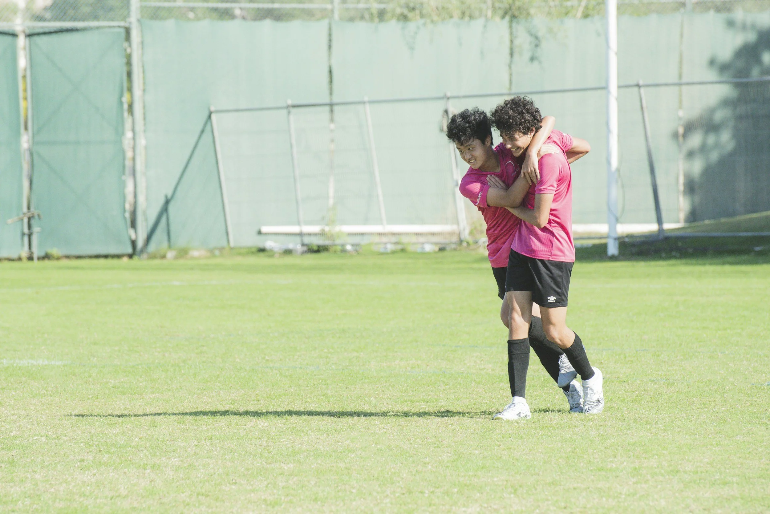 Two young women wearing pink sports shirts, black shorts, and athletic shoes are on a grassy sports field. One woman is carrying the other on her back. They are smiling and appear to be enjoying themselves. In the background, there is a green fence and some trees.