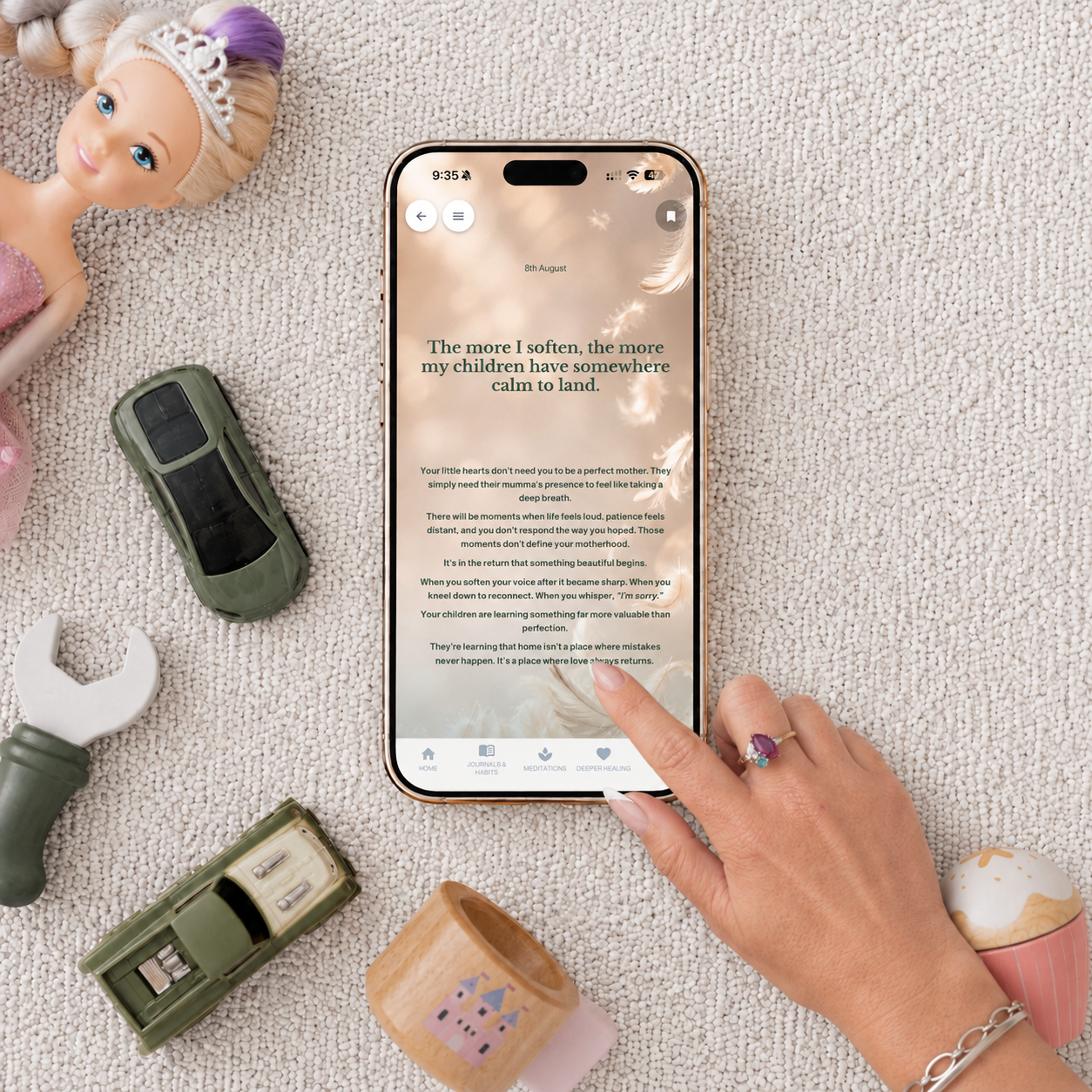 Smartphone on textured surface displaying inspirational text about motherhood with a hand touching the screen. Surrounding items include a doll with a tiara, toy cars, a wrench, a small candle holder with a castle design, and a decorative cupcake container.