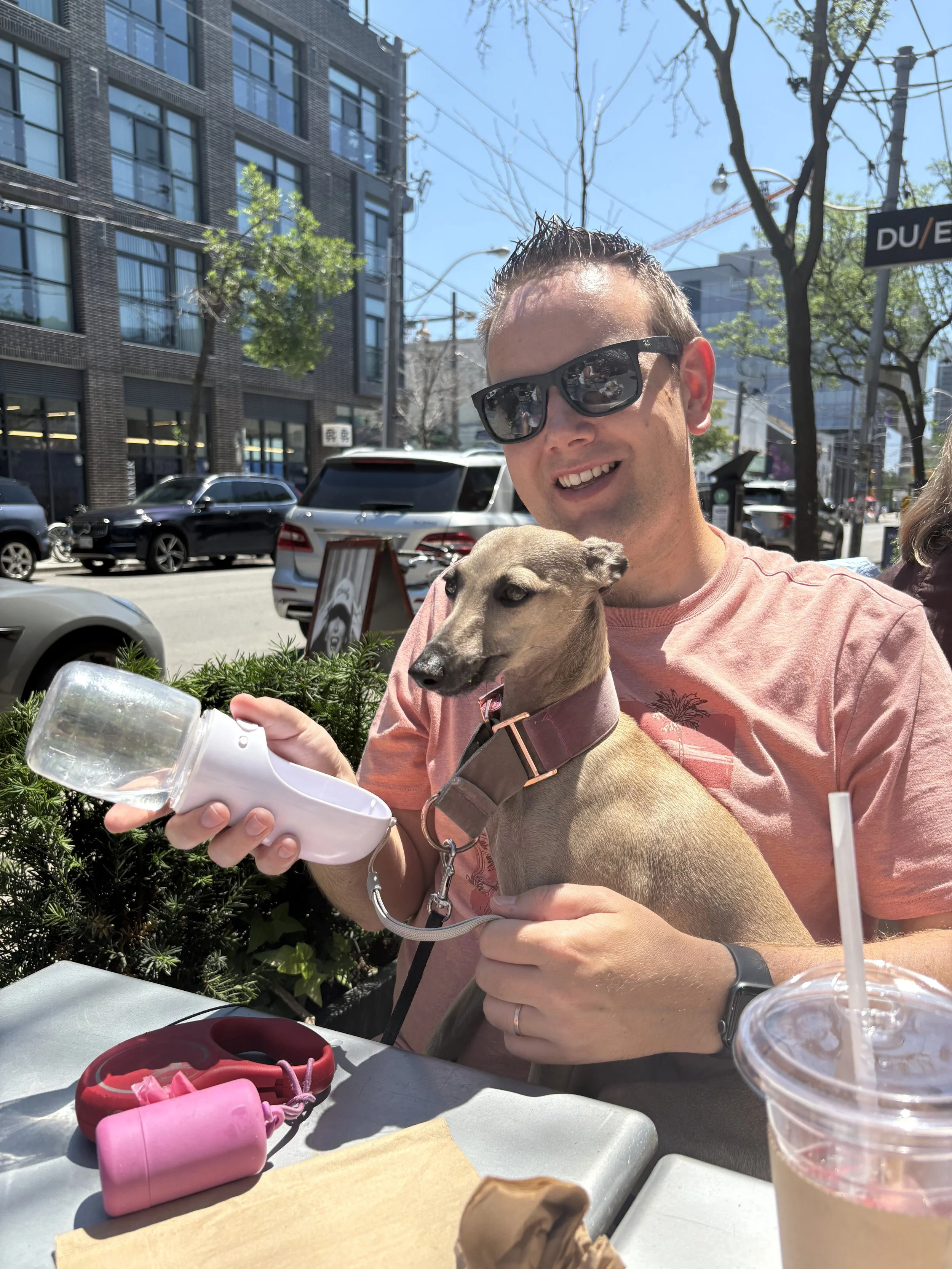 A man wearing sunglasses holds a small dog in his lap at an outdoor café table. The man is smiling and the dog appears relaxed, with a water bottle in the man's hand. There are cars parked along the street and buildings in the background.