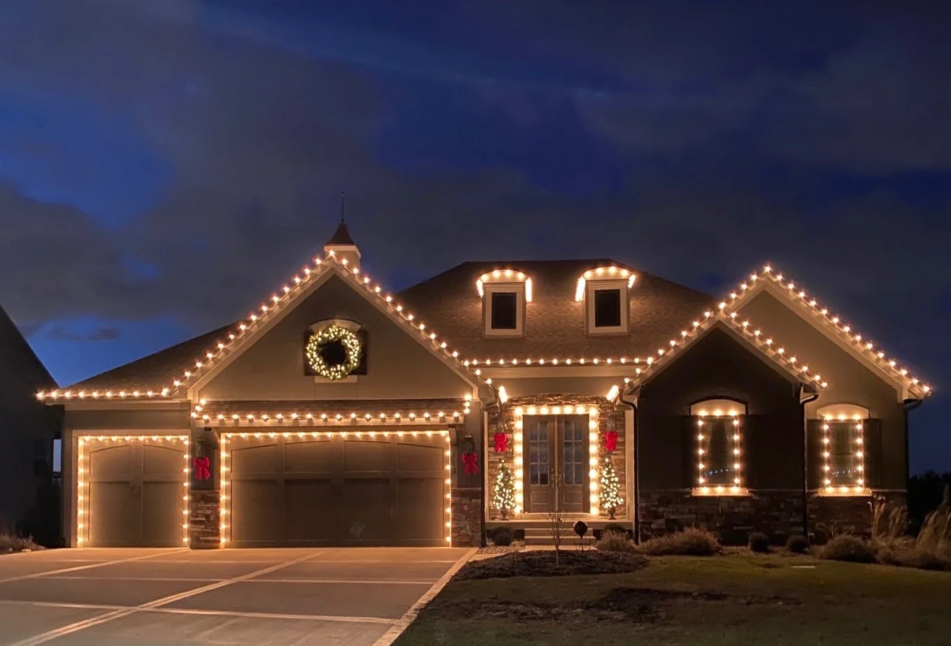 A house decorated with Christmas lights, including a large wreath and two small Christmas trees with lights, and red bows on the front porch, during dusk.