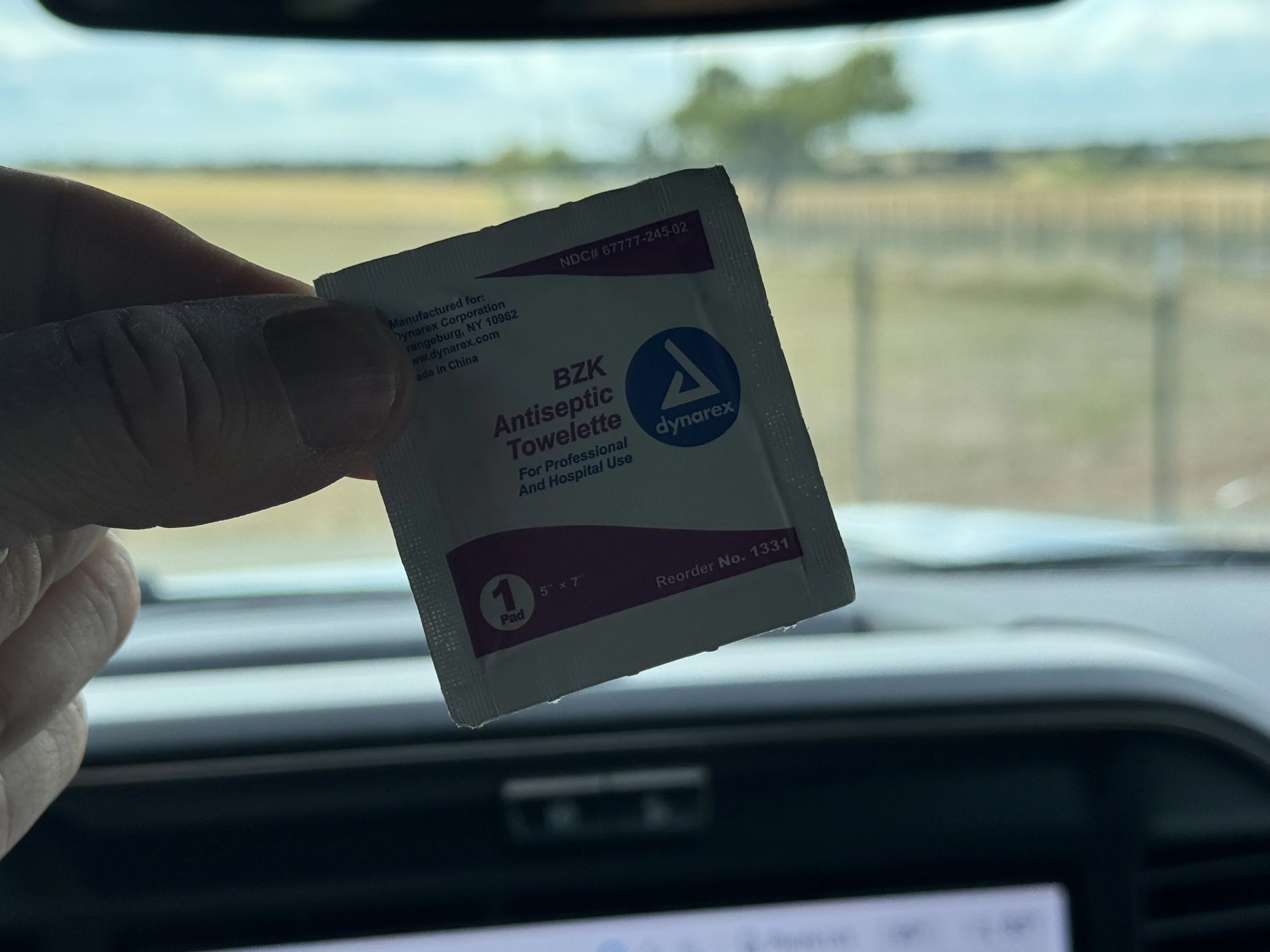 A hand holding a packet of antiseptic towelette in front of a vehicle's dashboard and windshield, with a blurred rural landscape outside.