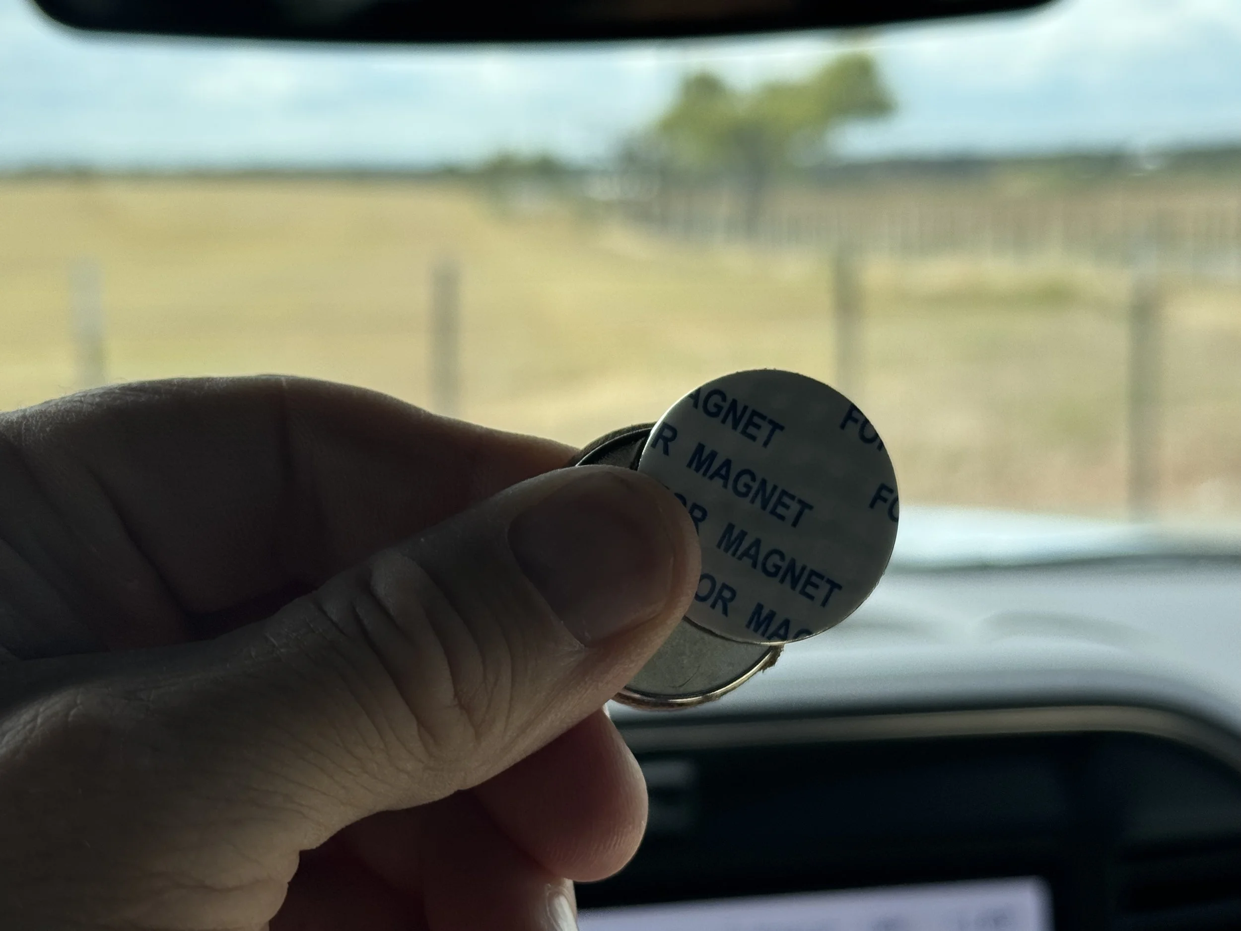 Hand holding a small round mirror with magnetic backing, labeled 'FOR MAGNET', inside a vehicle with a blurred outdoor landscape visible through the window.