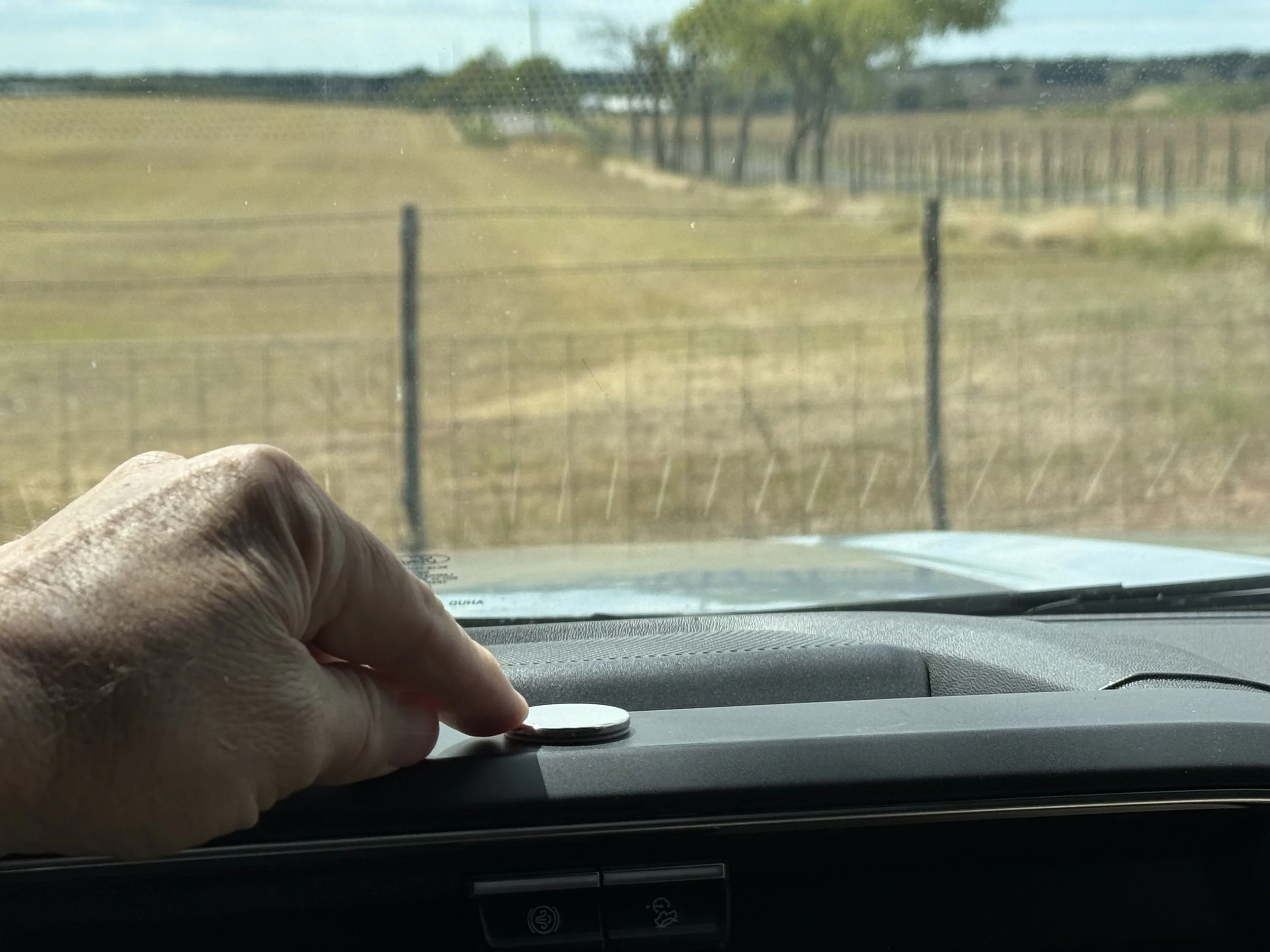 Inside a vehicle, a person's hand is placing a small metal disc on the dashboard near the windshield, with a rural landscape and a wire fence visible outside.