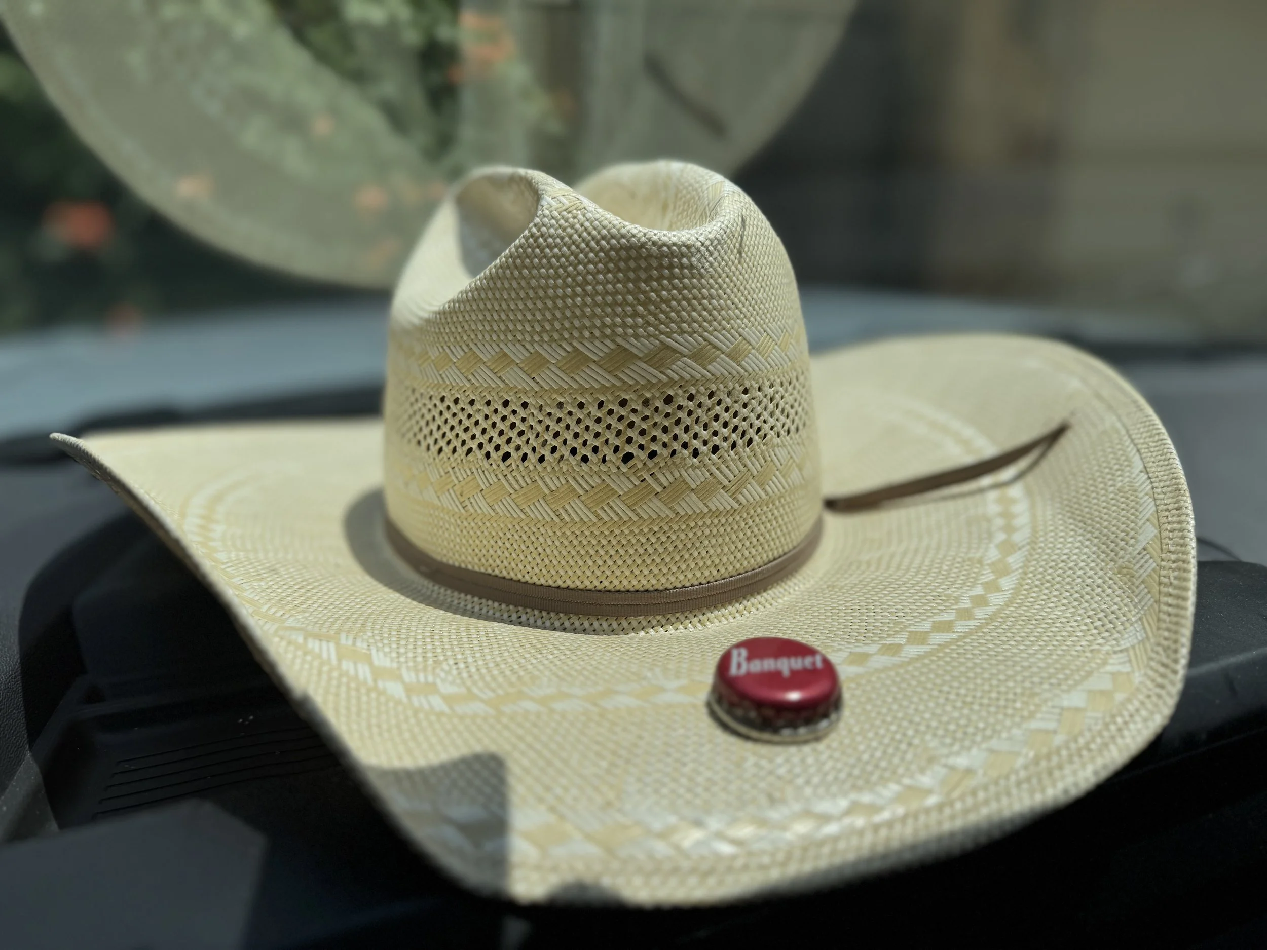 A straw cowboy hat with a decorative band and a red button that says 'Banquet' is placed on a car dashboard with a blurred background.