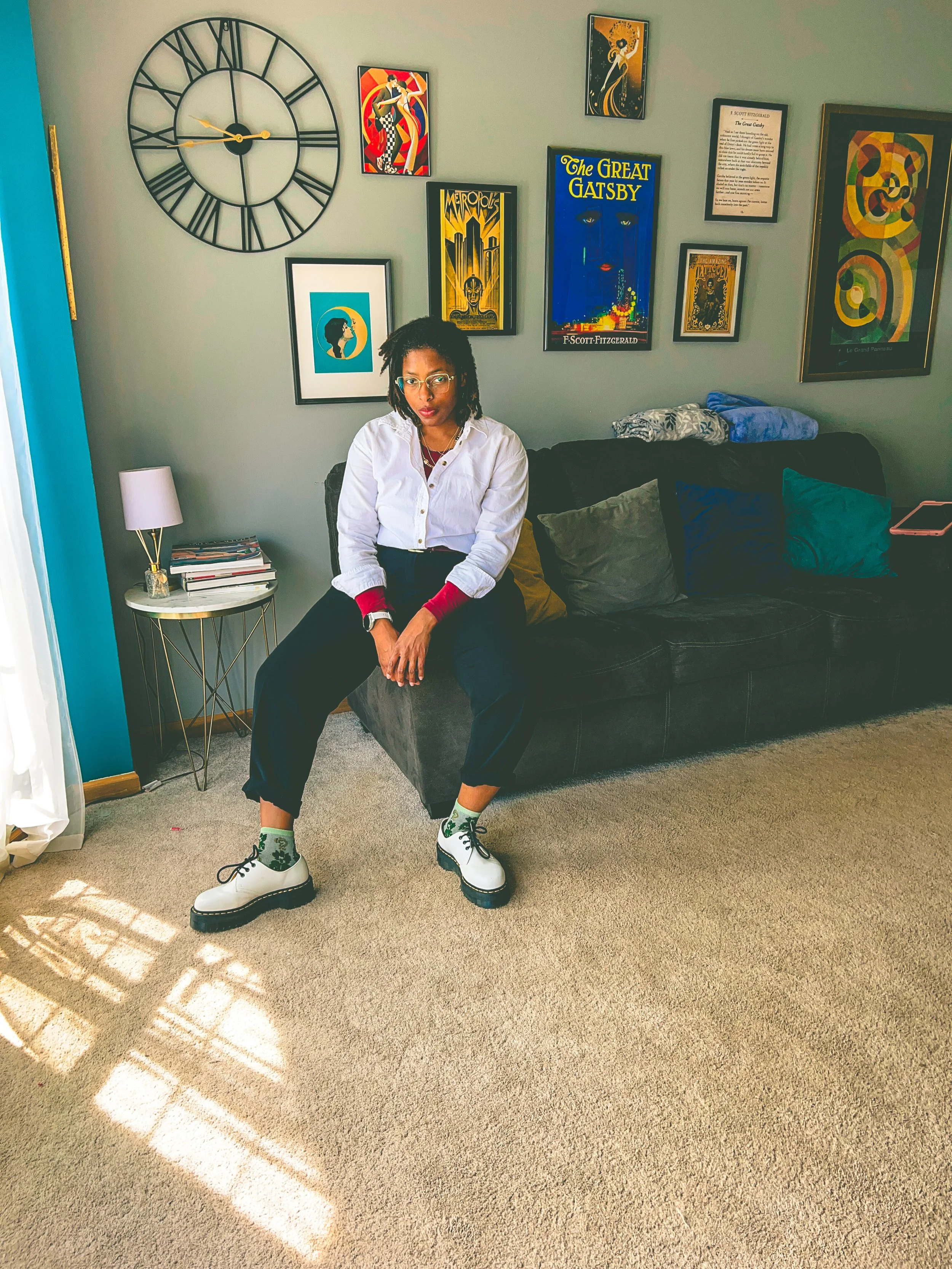 A woman with glasses sitting on a black couch in a living room, with a gallery wall of colorful artwork including a large clock behind her, sunlight casting shadow patterns on the beige carpet.