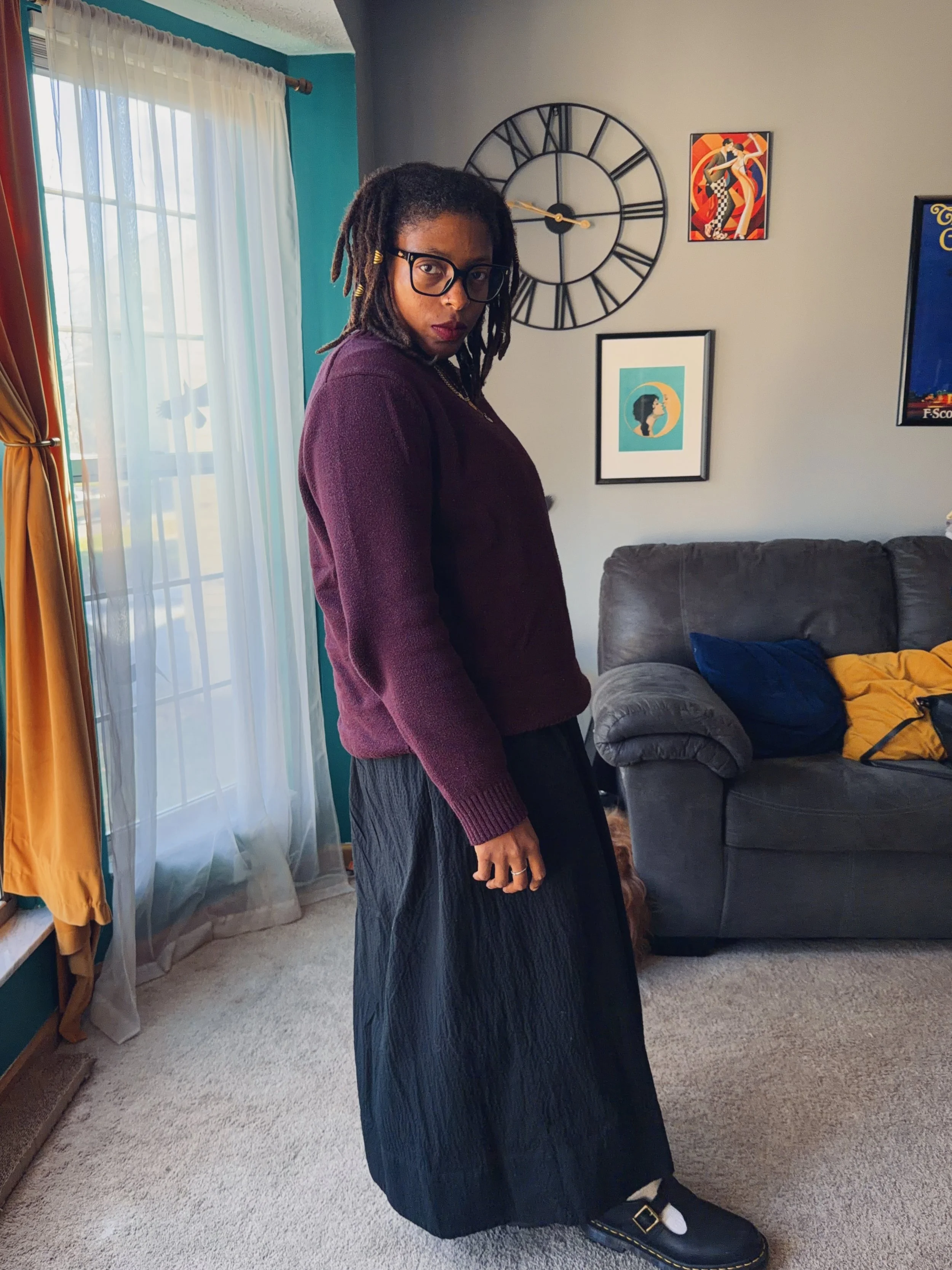Woman with glasses and dreadlocks standing in a living room near a gray couch, with colorful artwork and a wall clock behind her, sunlight coming through a window with sheer curtains.