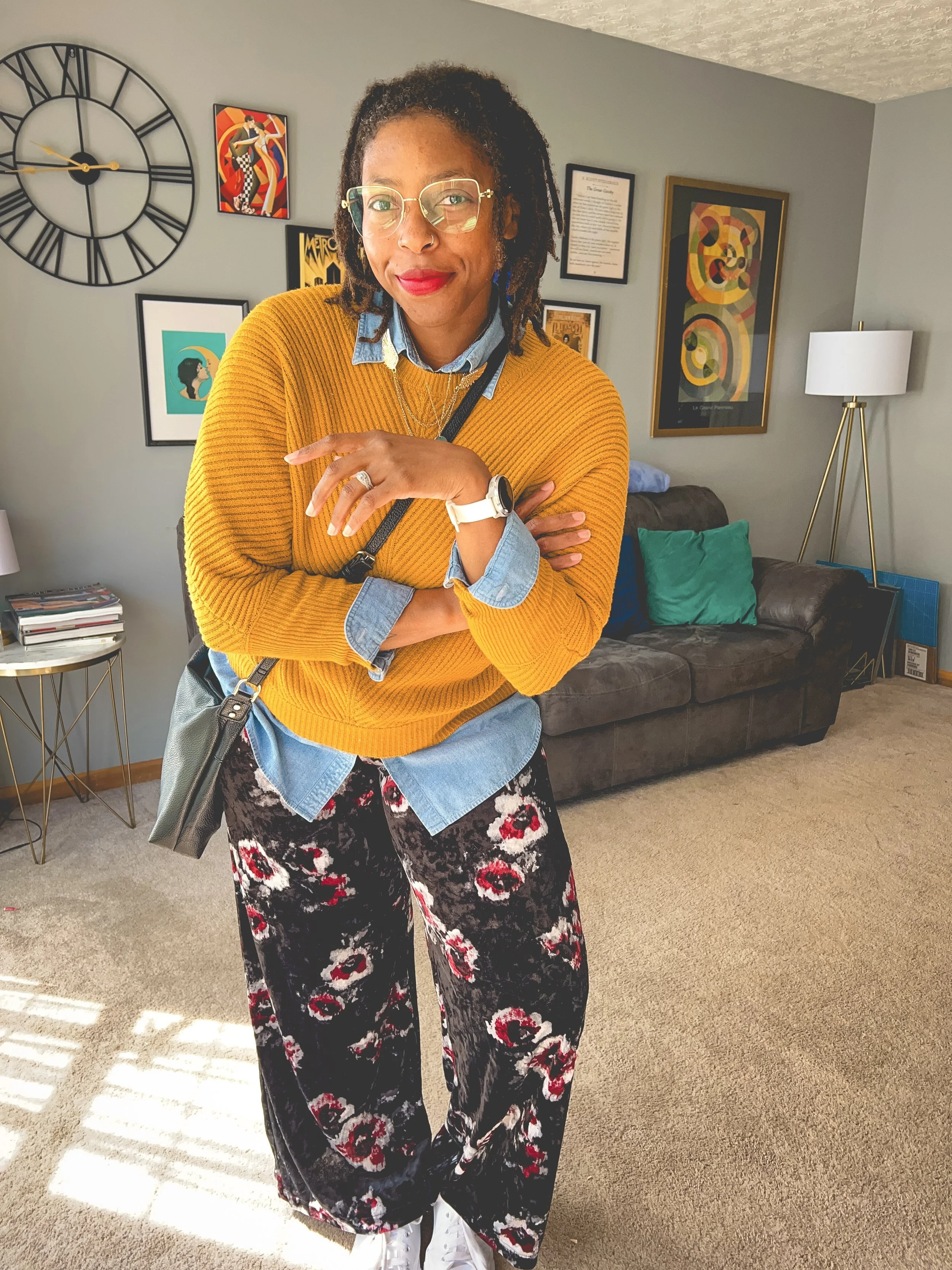 A woman with glasses and red lipstick standing in a living room, wearing a yellow sweater layered over a chambray shirt, patterned black and red pants, and white sneakers, with her arms crossed.