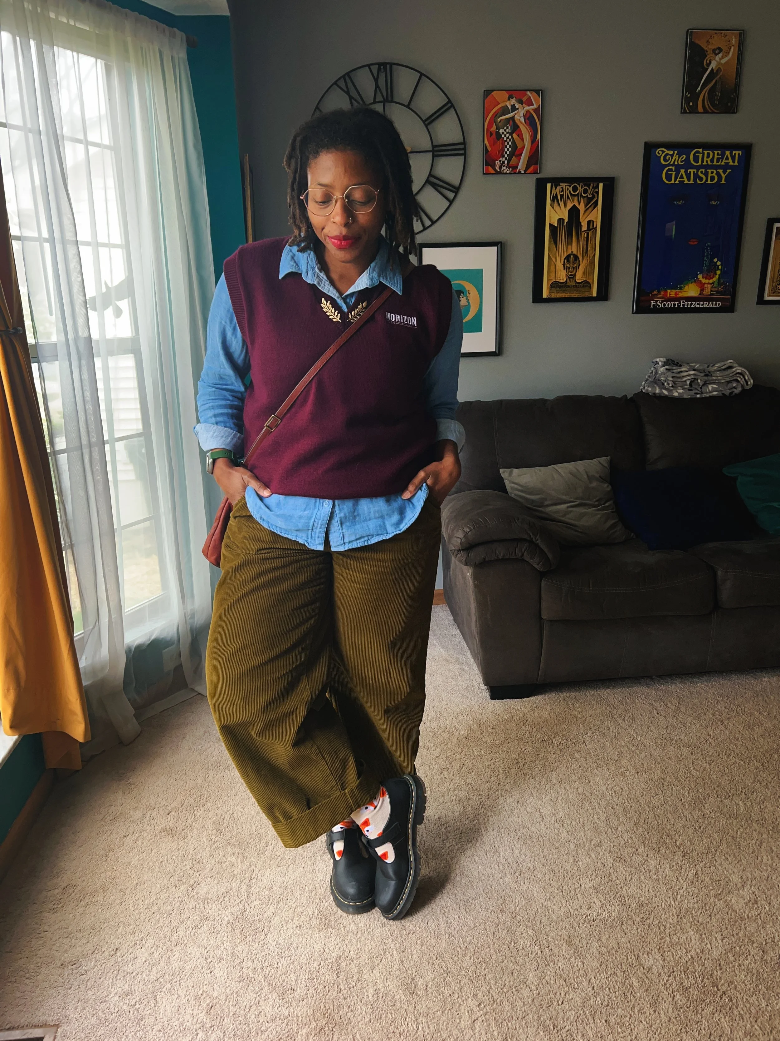 A woman standing indoors with her hands in her pockets, wearing glasses, a blue shirt, a maroon vest, olive green pants, colorful socks, and black boots, with artwork and a large clock on the wall behind her.