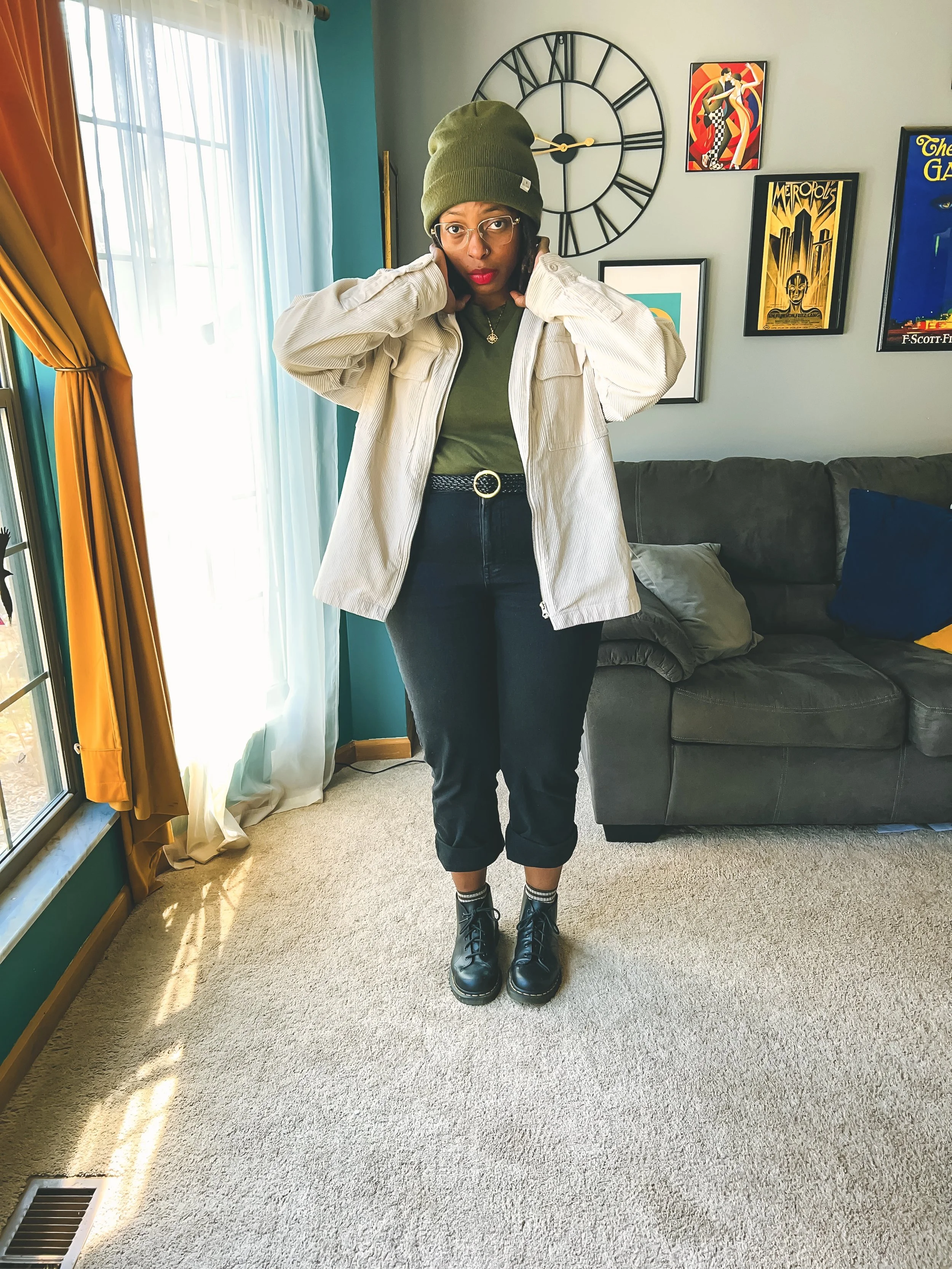 A woman standing indoors, wearing glasses, a green beanie, a beige jacket, green shirt, black pants, and black combat boots, with a large wall clock and framed posters on the wall behind her.
